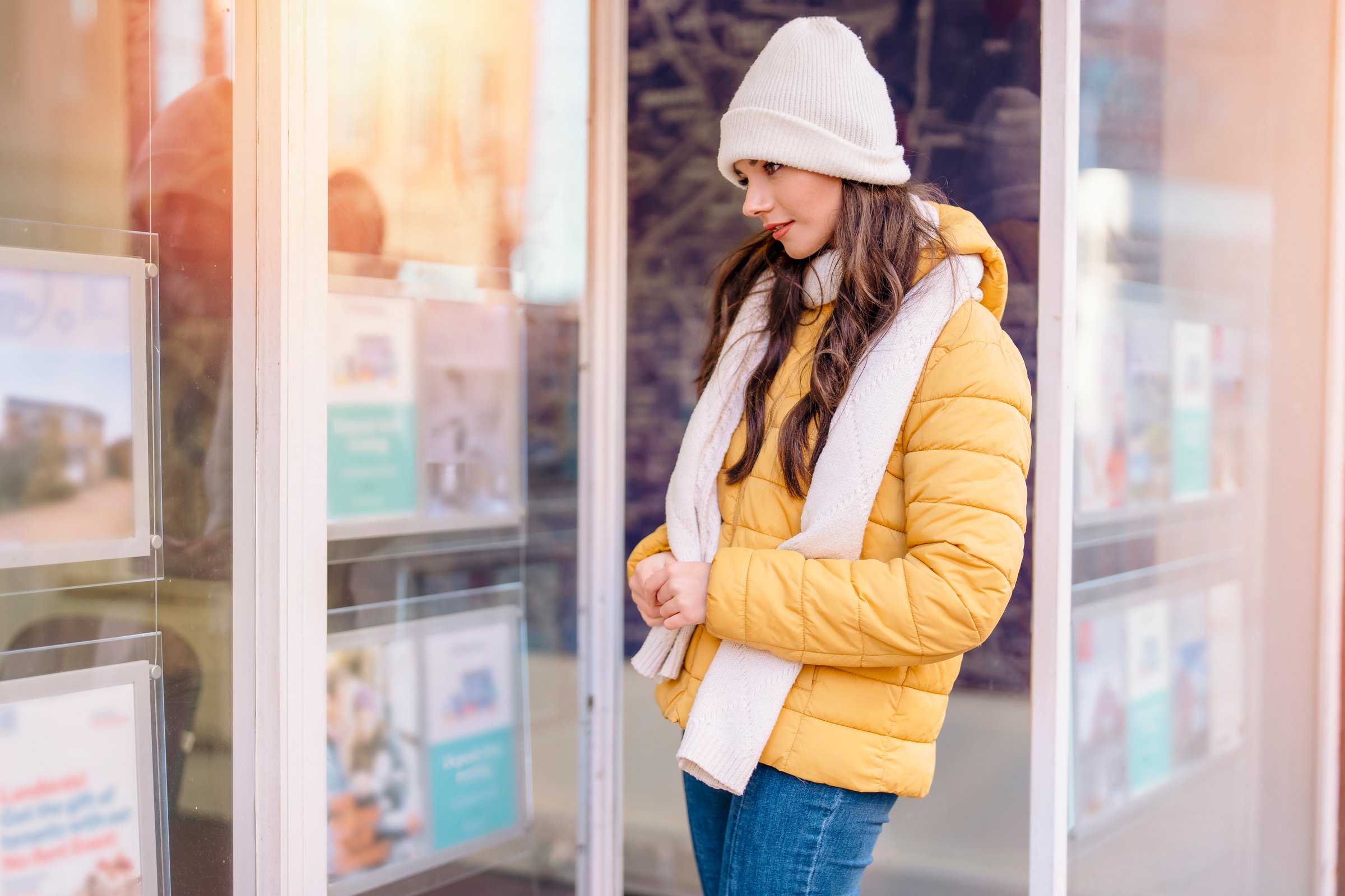 Woman in yellow jacket and white beanie looking at real estate listings in a window display on a sunny day.
