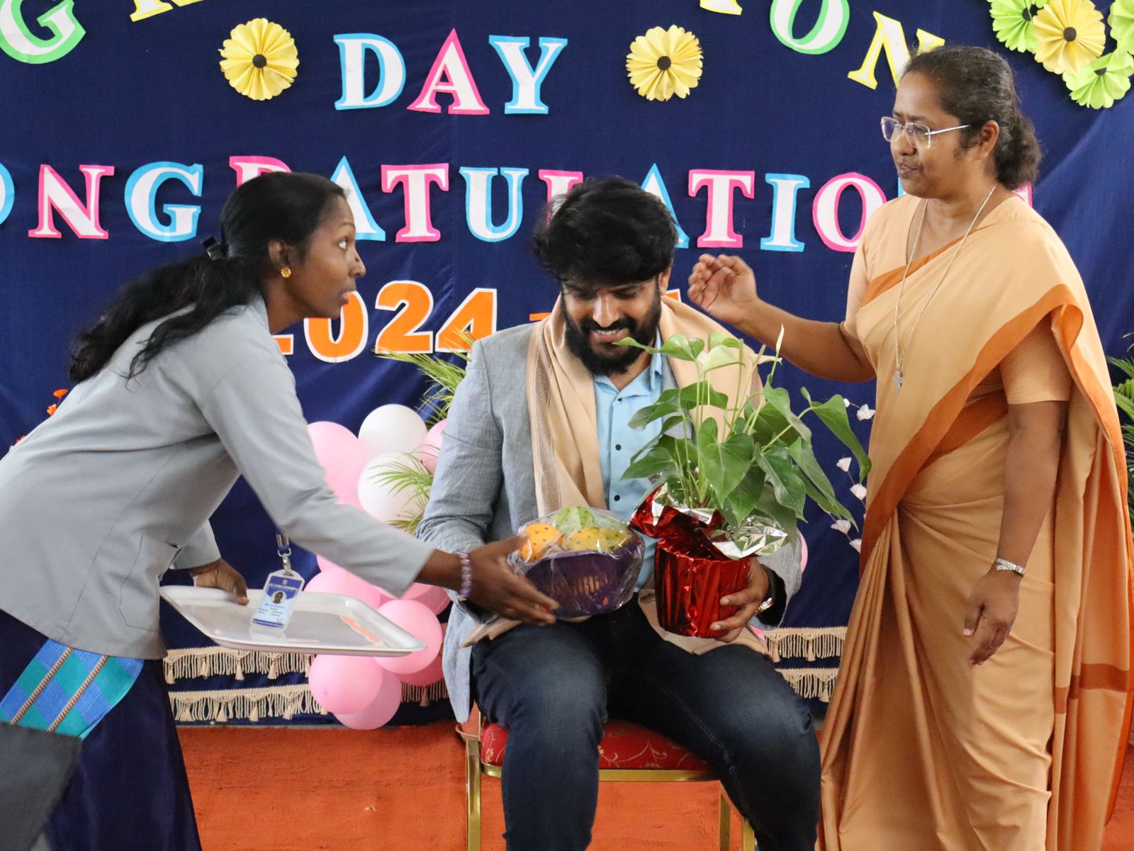 Man receives bouquet and plant from two women at a celebration event with congratulatory banner in the background.