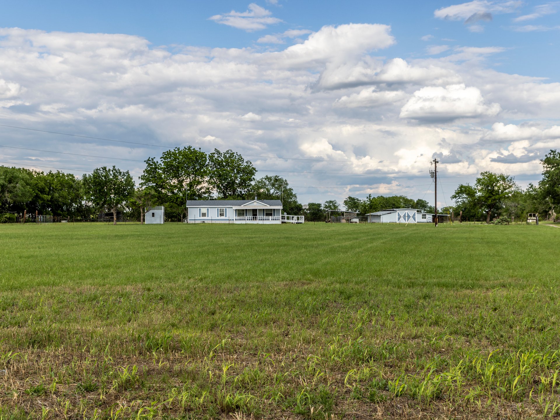 A white house with a porch in a vast green field under a cloudy sky, surrounded by trees and a utility pole.