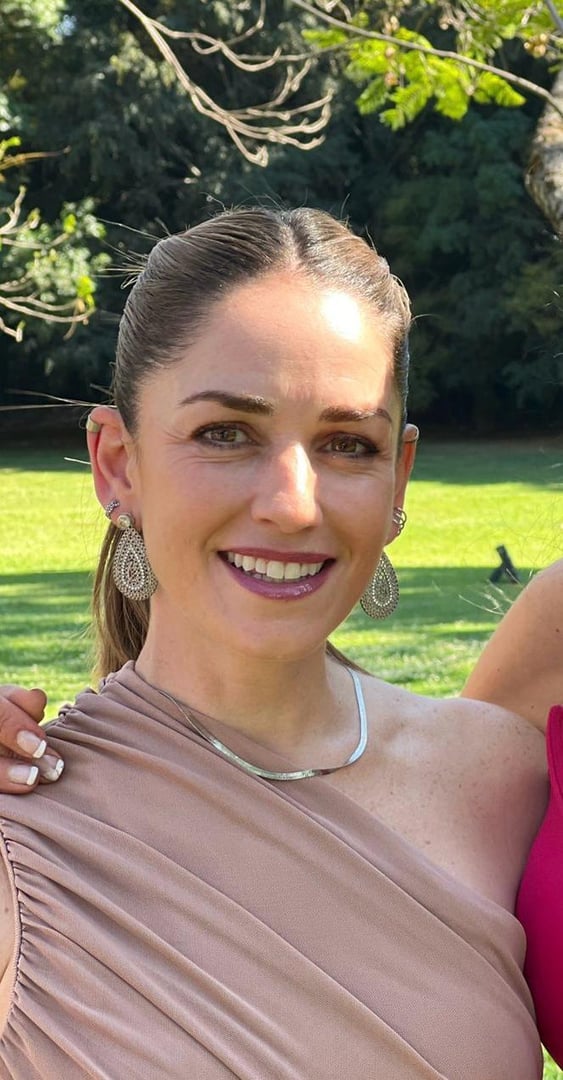 Woman outdoors wearing a one-shoulder dress, silver necklace, and large earrings, smiling with trees in the background.