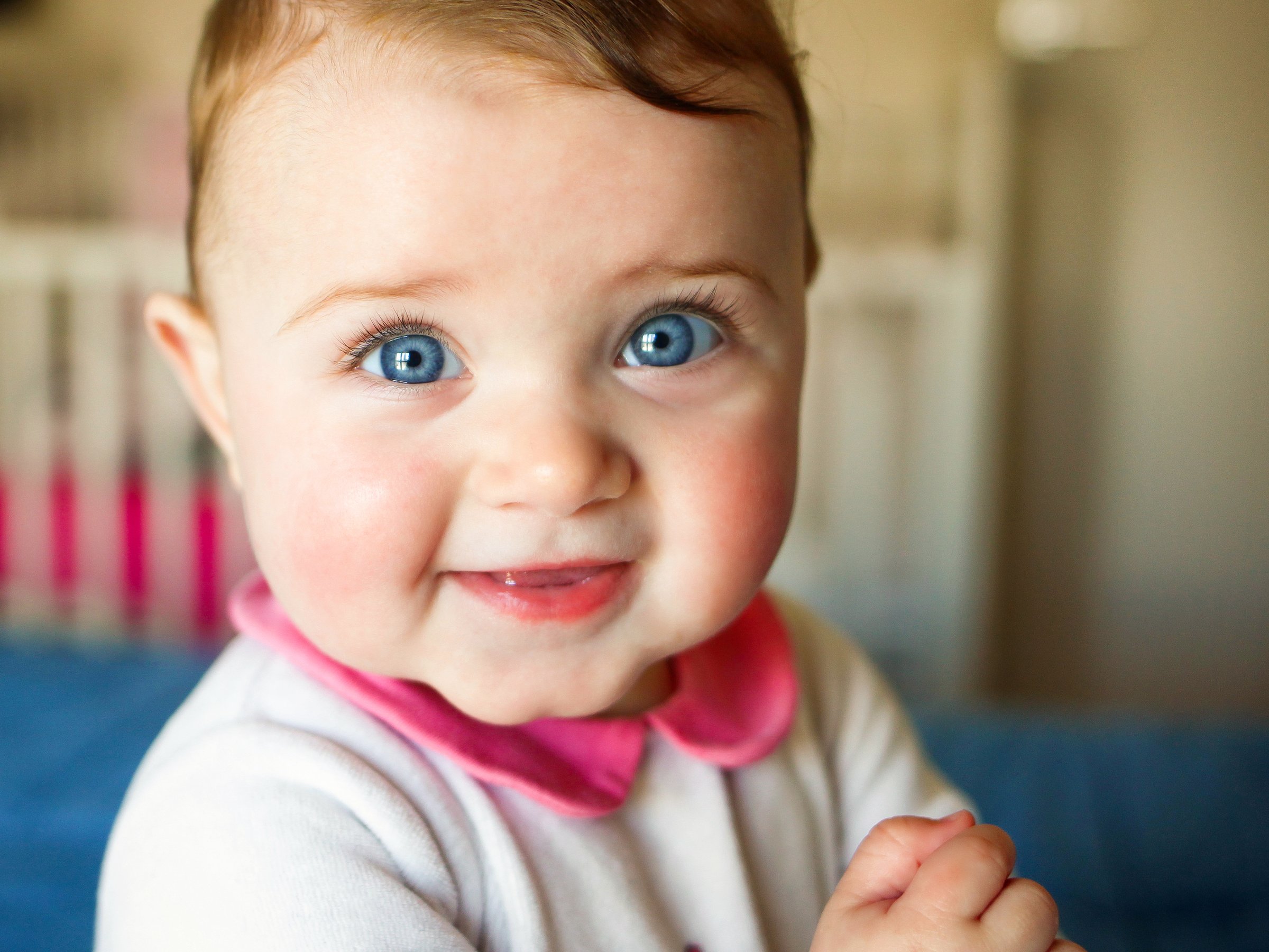 A smiling baby with bright blue eyes, wearing a pink-collared white shirt, and clasping hands, in a softly blurred background.