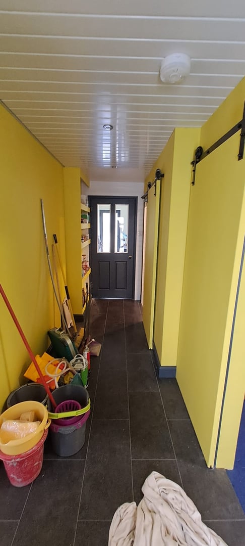 Hallway with yellow walls, tile floor, sliding doors, and cleaning supplies in buckets and mops on the left side.