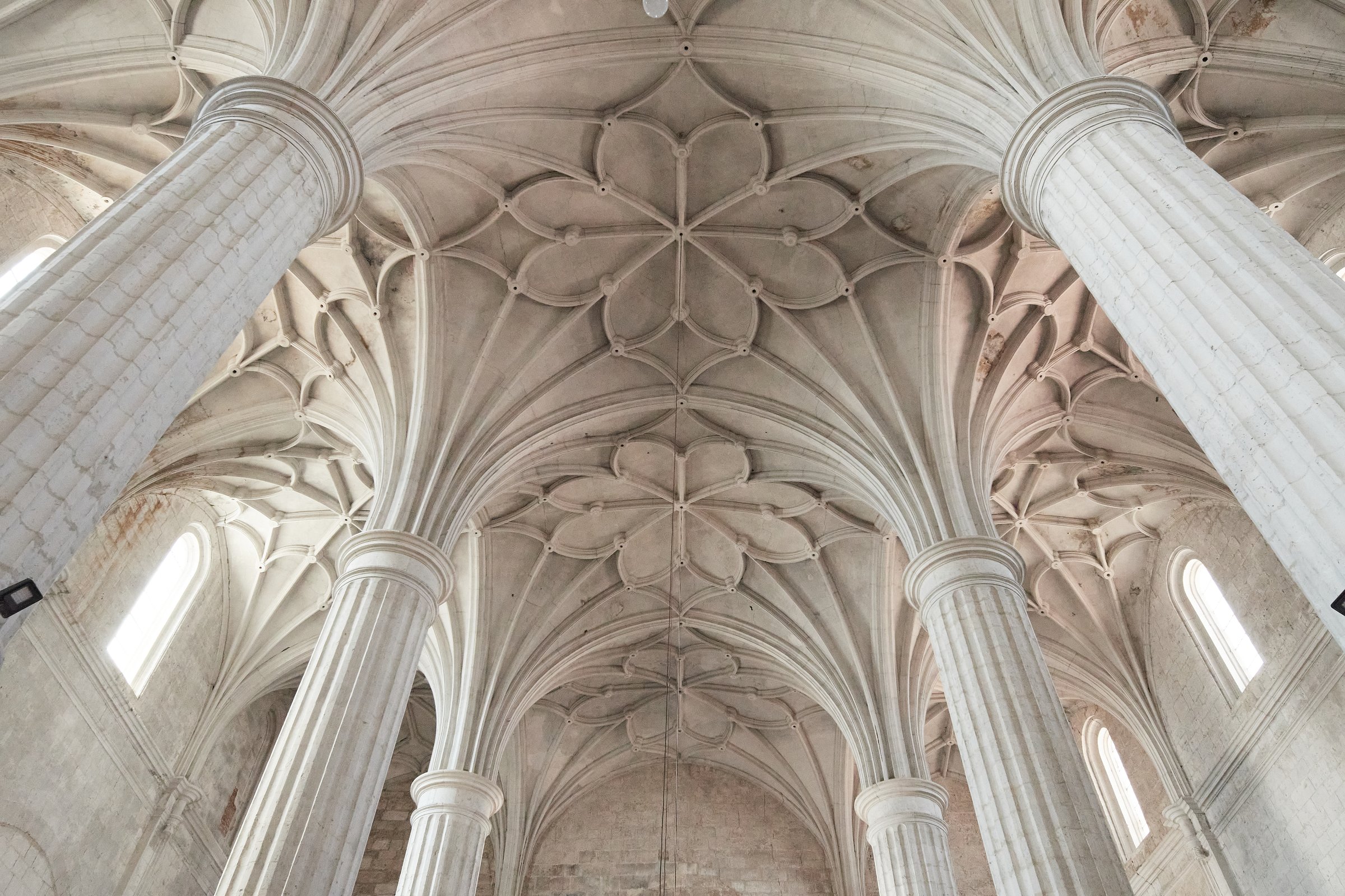 The image captures the stunning intricate details of a vaulted ceiling supported by grand stone columns inside a historic church. The architectural design showcases exquisite craftsmanship with its elaborate patterns and structural elegance. The high arches and symmetrical designs create a sense of awe and reverence, highlighting the historical and cultural significance of the building. The natural light streaming through the windows enhances the beauty of the stonework, emphasizing the grandeur and timelessness of this sacred space.