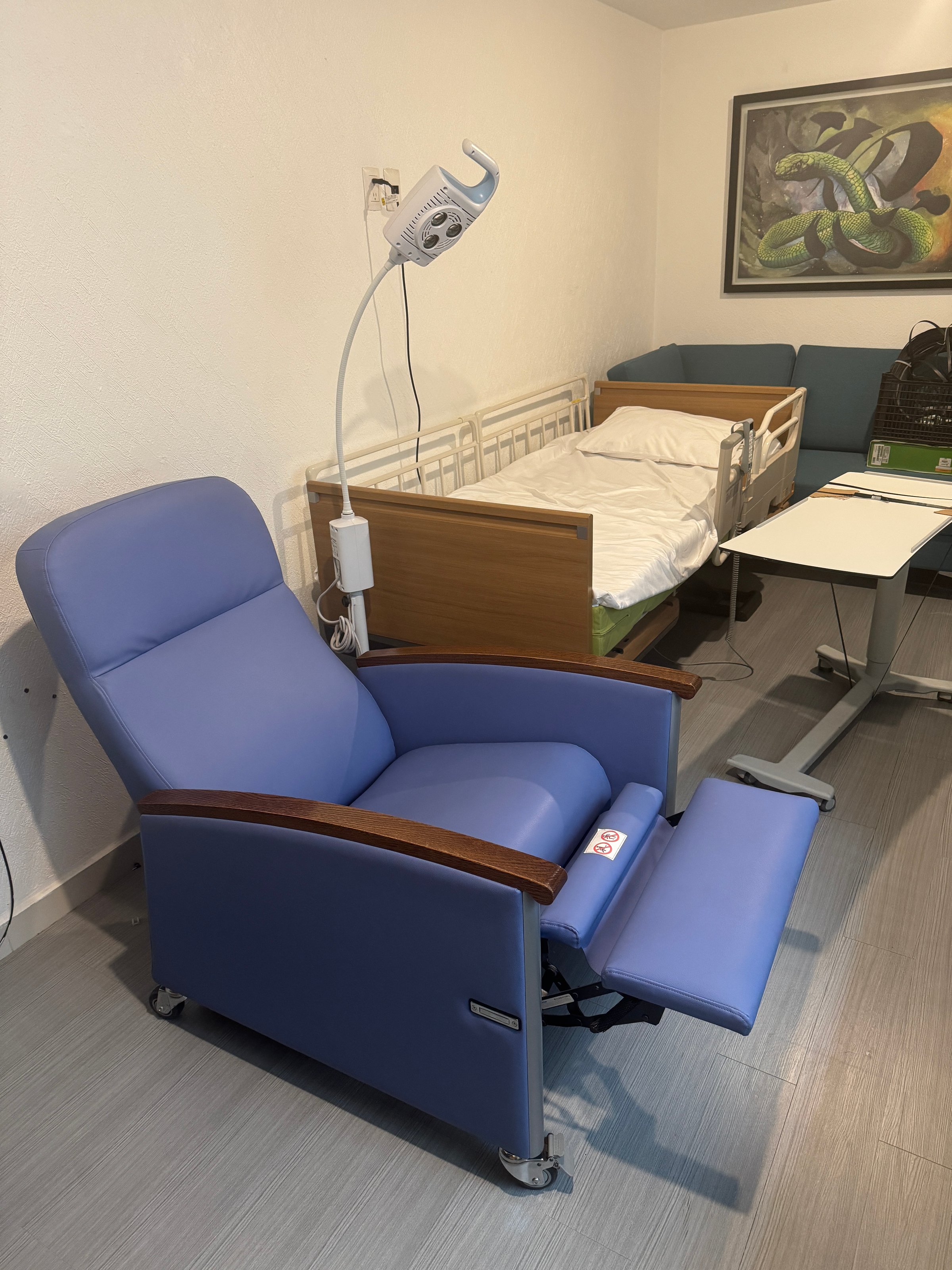 Hospital room with a blue reclining chair, hospital bed, and adjustable overbed table on a light grey floor.