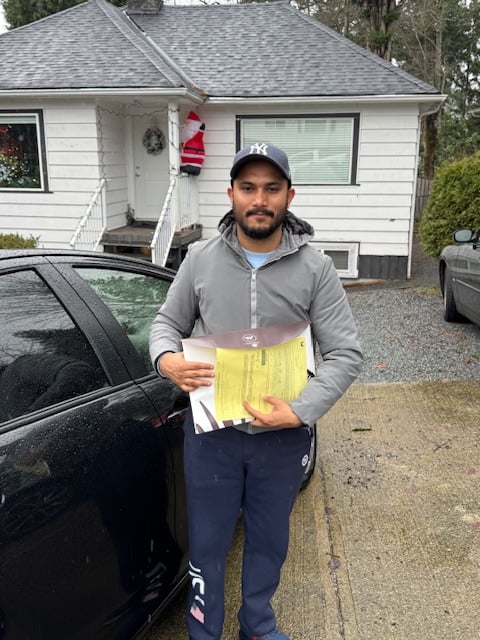 Person standing outside a house, holding papers, next to a car on a rainy day.