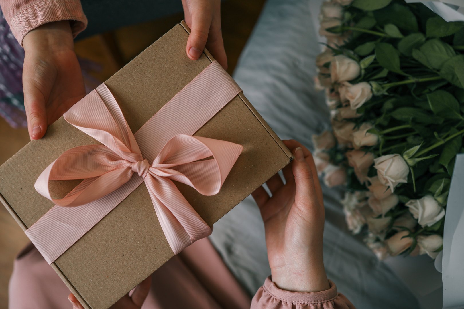 Hands of a child giving a wrapped gift with a satin ribbon to a woman holding a bouquet of roses, captured in warm indoor light.