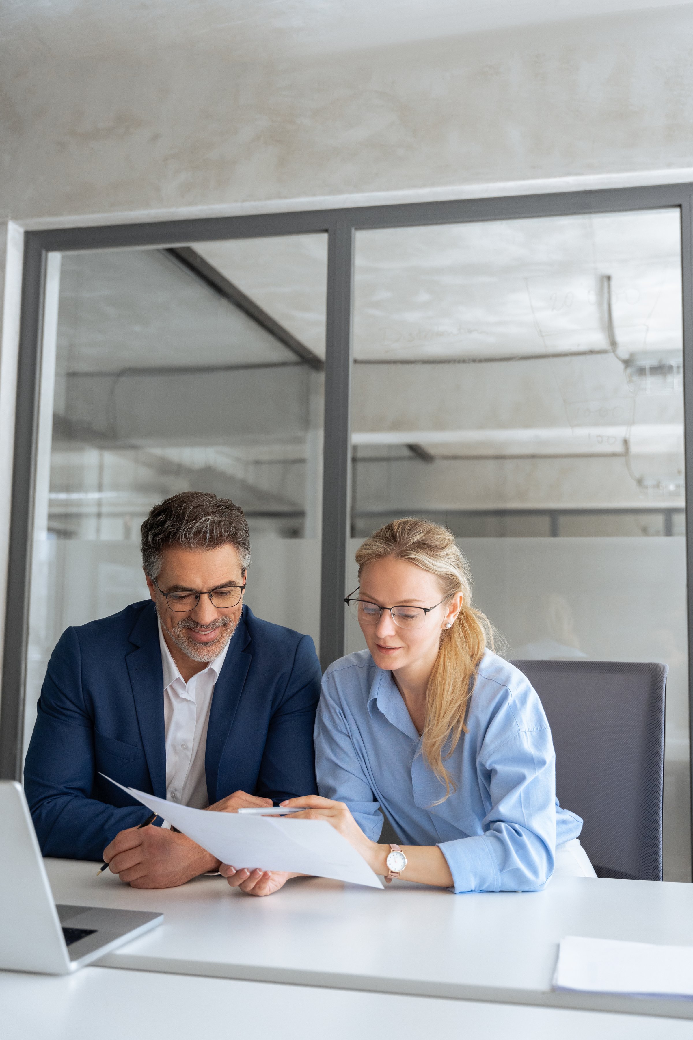 Two professional business people working together. Partners mature man and woman discussing financial document report sitting at table in office. Team leader explain trainee working process. Vertical