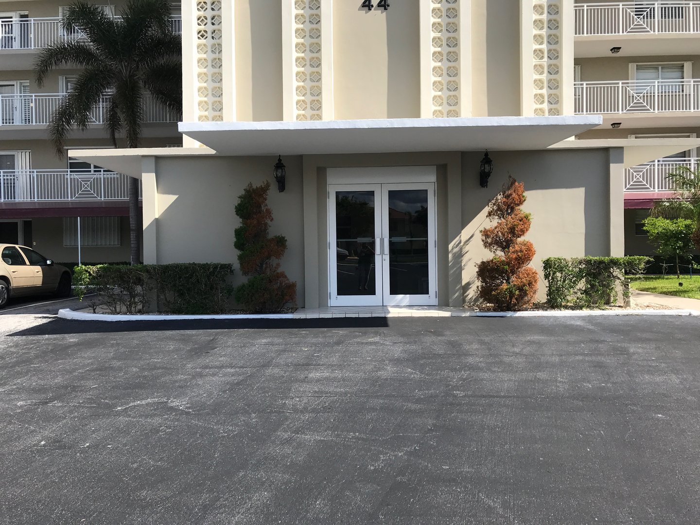 Entrance of a beige building with glass double doors, surrounded by trimmed shrubs and a paved driveway.