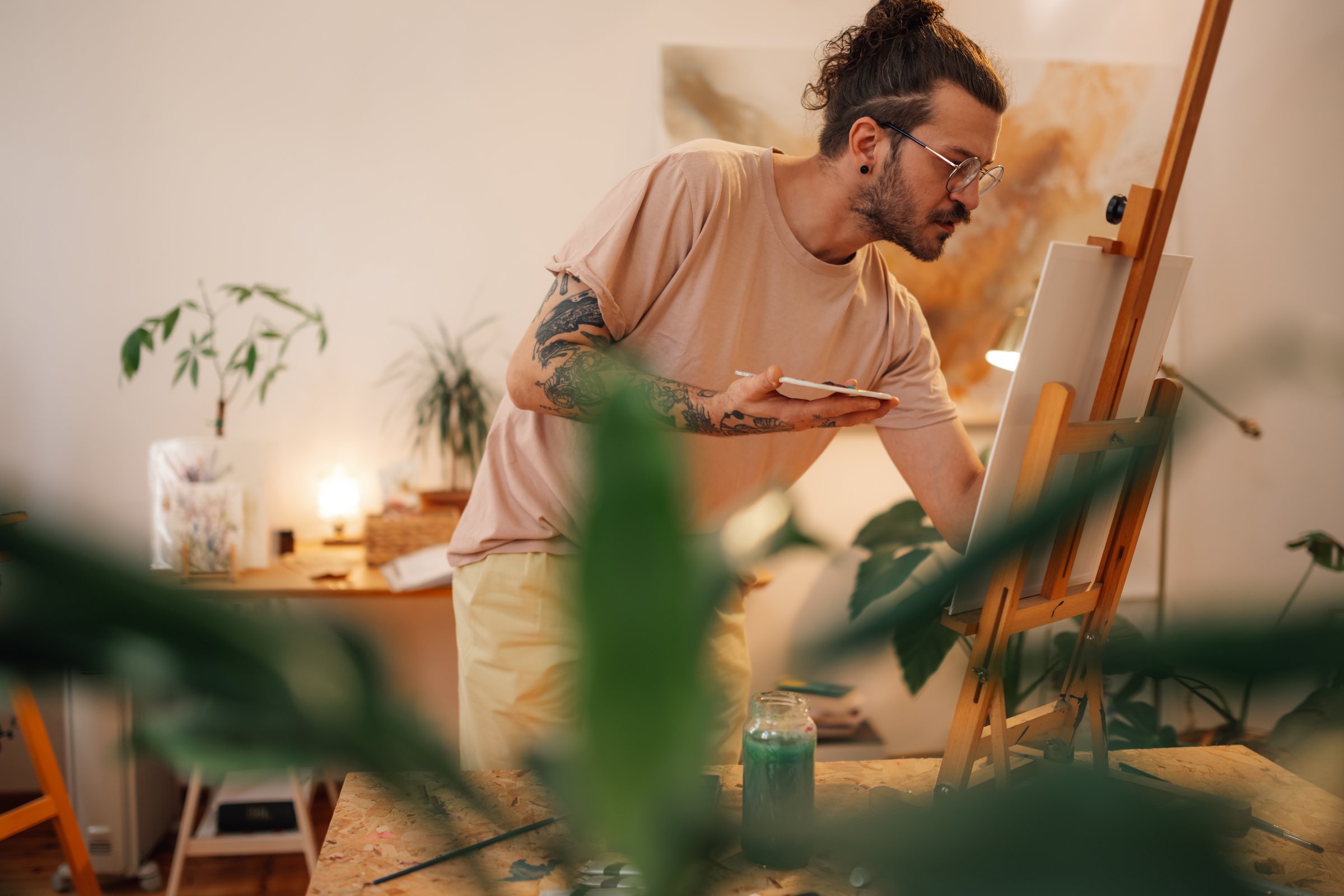 Portrait of tattooed trendy hipster artist standing at creative art studio with brush and palette in hands and painting on easel. Precise focused young painter at atelier and painting on canvas.