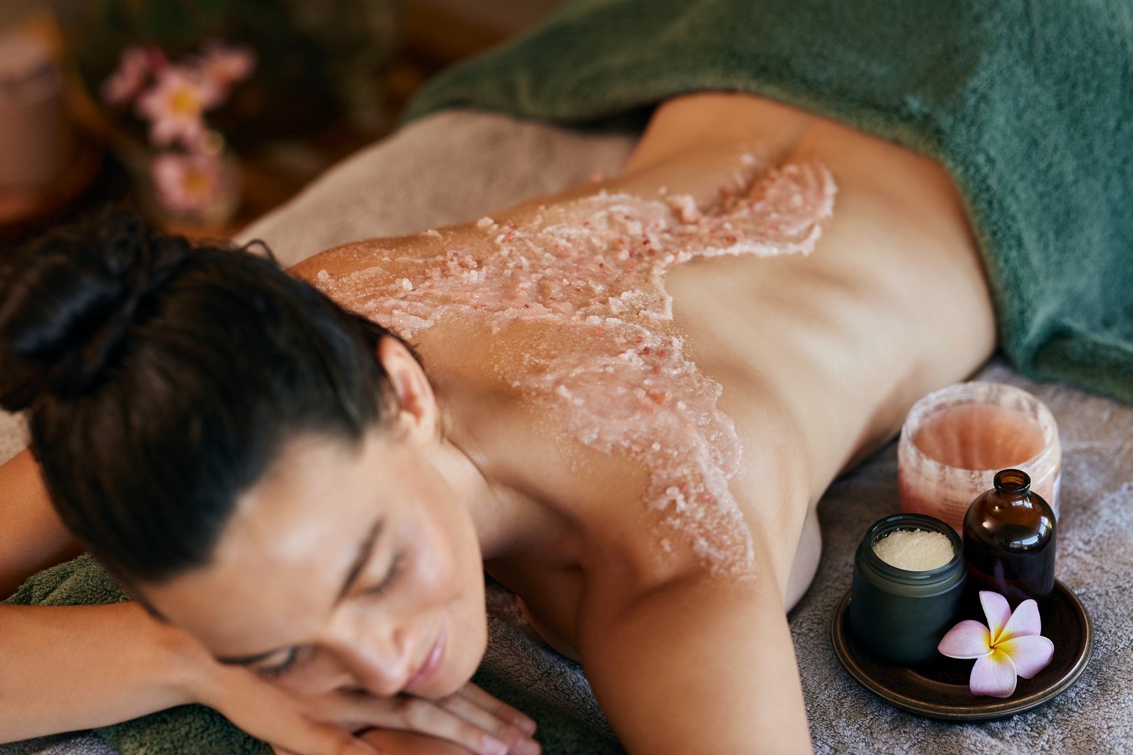 Woman lying down on massage table while relaxing during exfoliating salt body scrub applied on back. Spa therapist using pink Himalayan salt scrub with aroma and essential oils in rejuvenation center. Woman feeling relaxed during spa treatment.