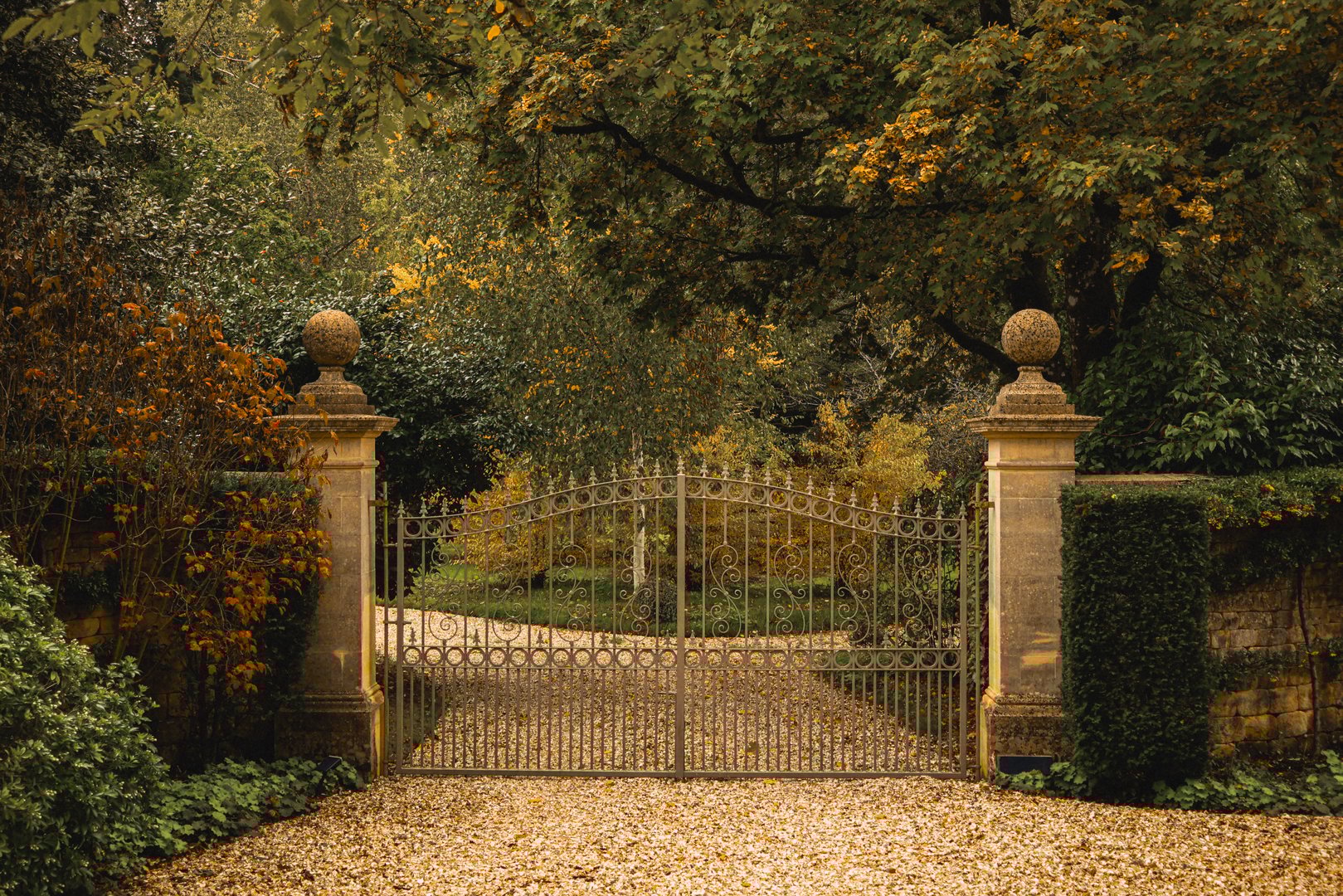 Grand wrought iron gate standing between stone pillars, leading to an elegant estate driveway in autumn. Cotswolds countryside, England
