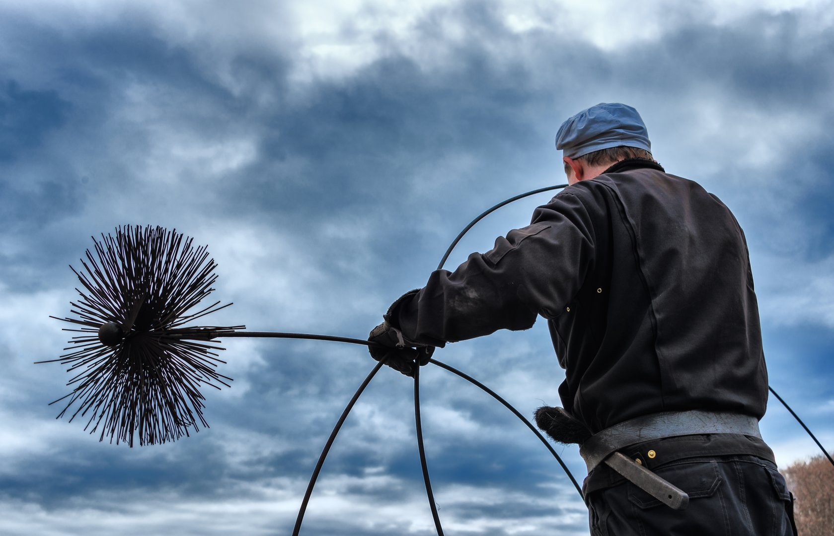 close up of a chimney sweep on the chimney at work; clad in the traditional workwear and holding a rope with the broom inside of the chimney;  dramatic sky in the background