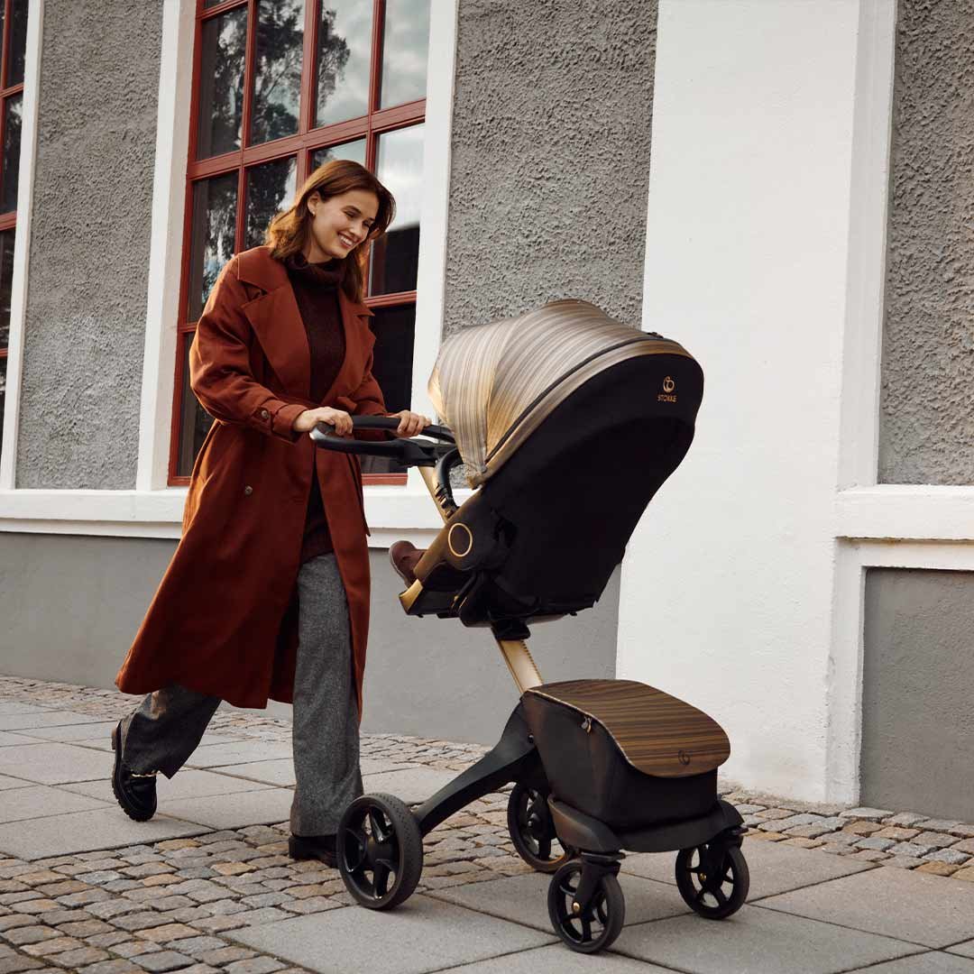 Person in a brown coat pushing a modern black and gold stroller on a sidewalk near a building with large windows.