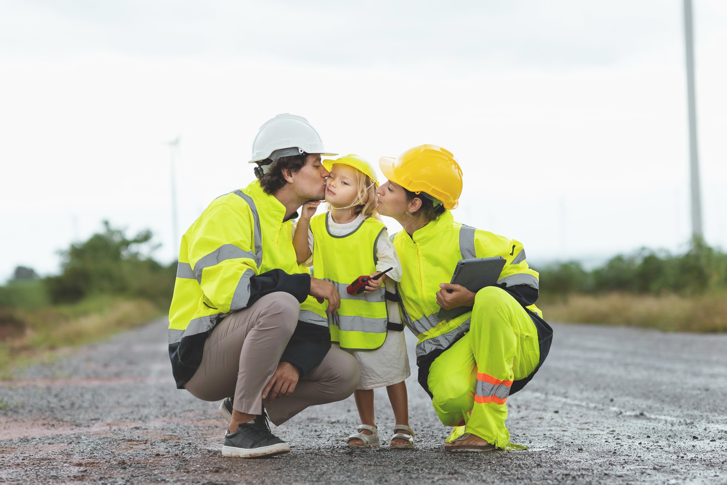 family of three, a man, a woman and a child, are standing on a road. The man is kissing the child on the cheek. The woman is holding a tablet. Scene is warm and affectionate