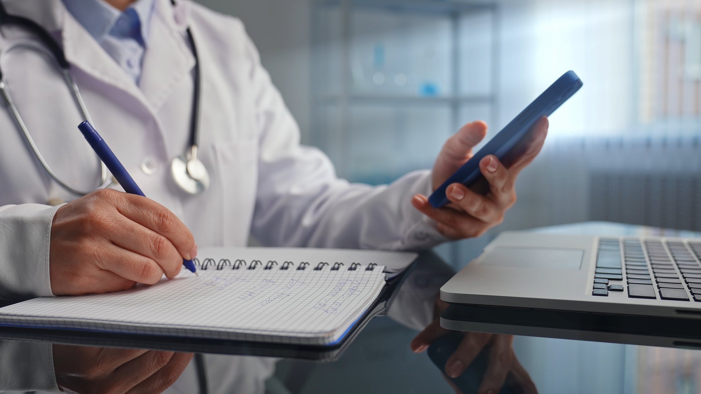 A doctor in a white coat is taking notes in a notebook while checking information on a smartphone and using a laptop.
