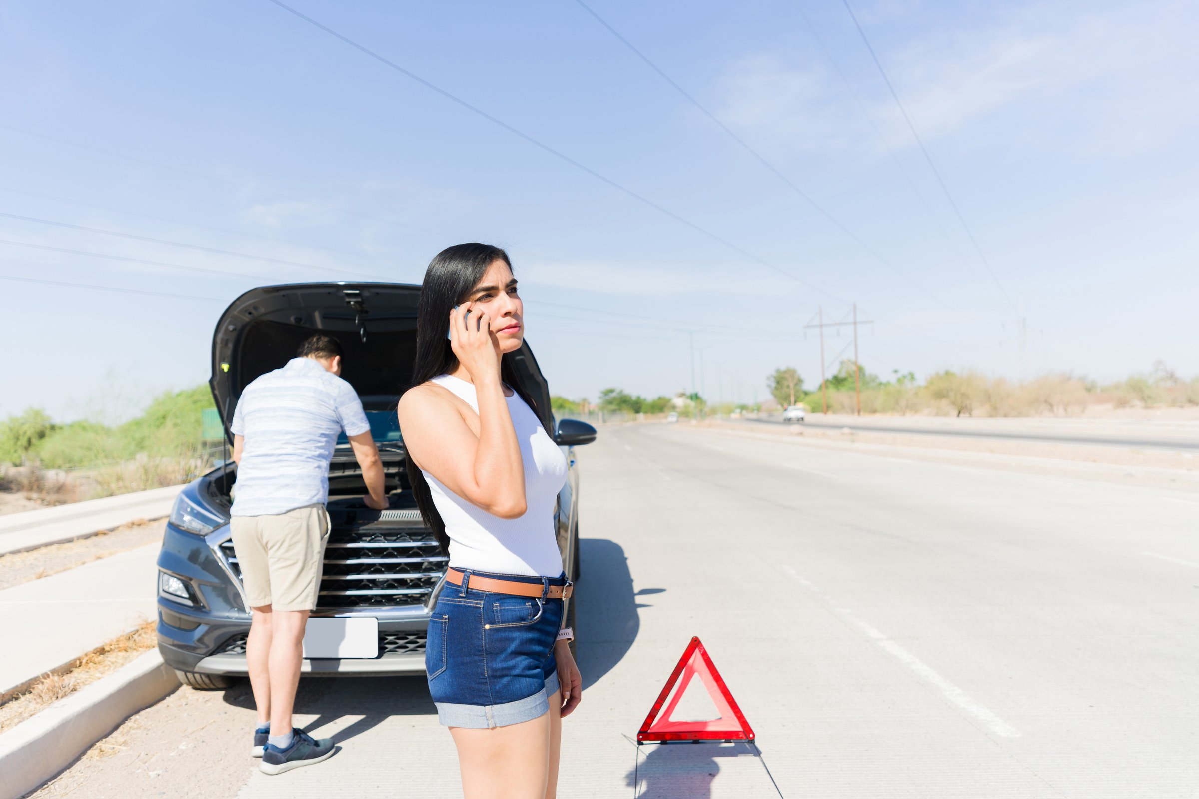Couple experiencing car trouble on a highway, man inspecting engine and woman calling for roadside assistance