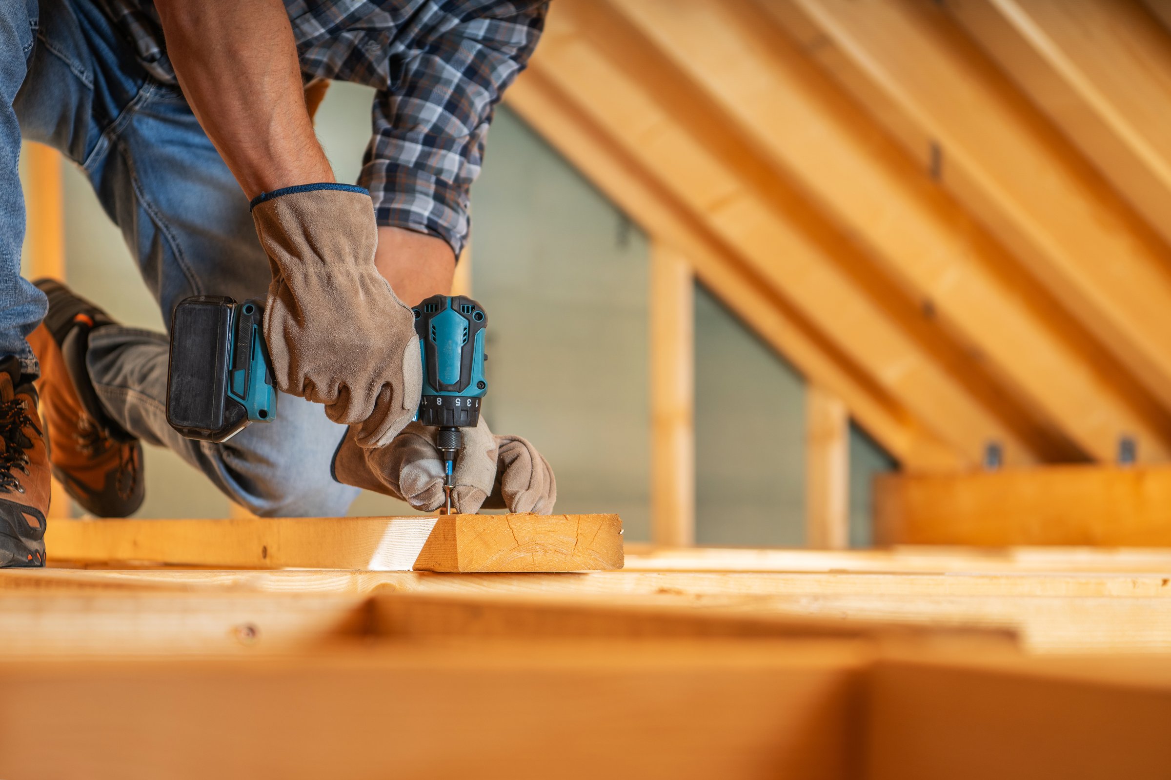 A skilled carpenter works diligently in an attic, securing wooden beams with a power drill while wearing gloves. The ambient light enhances the craftsmanship.