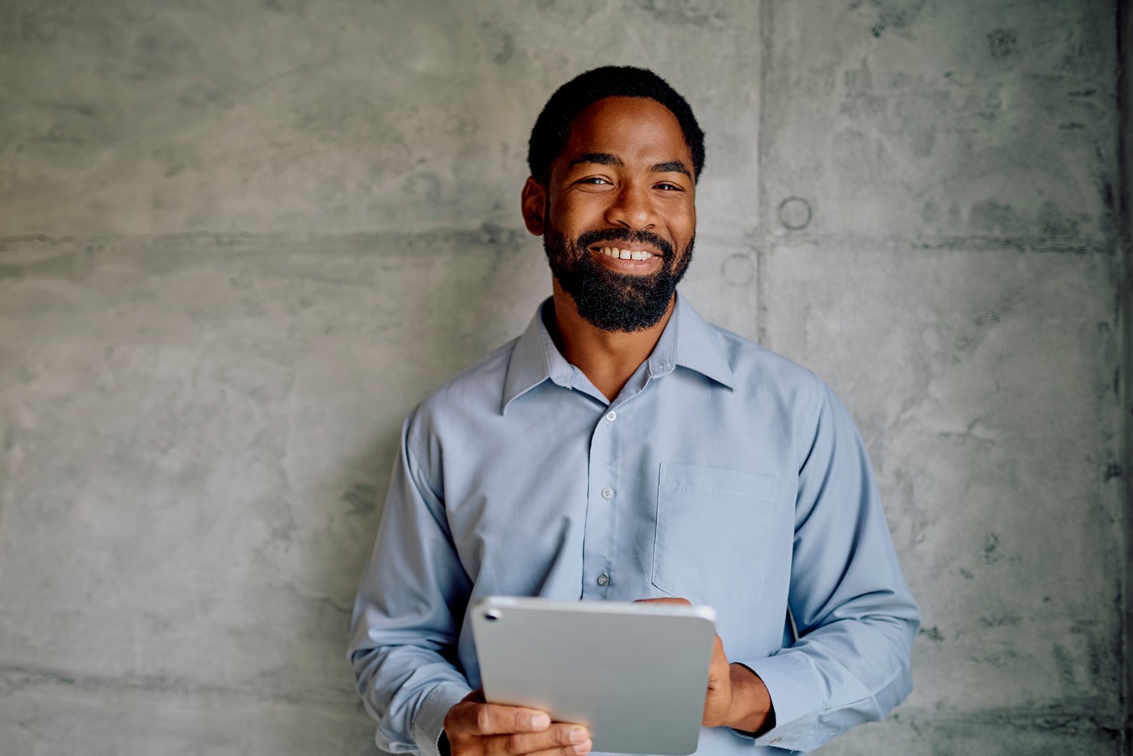 African American businessman smiling at camera, holding a digital tablet against concrete wall, representing modern business and technology