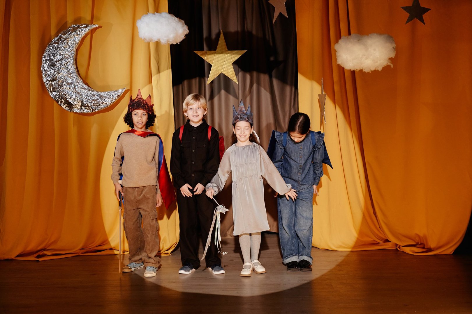 Young children standing on stage during a performance, holding hands and wearing costumes in front of a backdrop featuring stars and a crescent moon hanged from ceiling