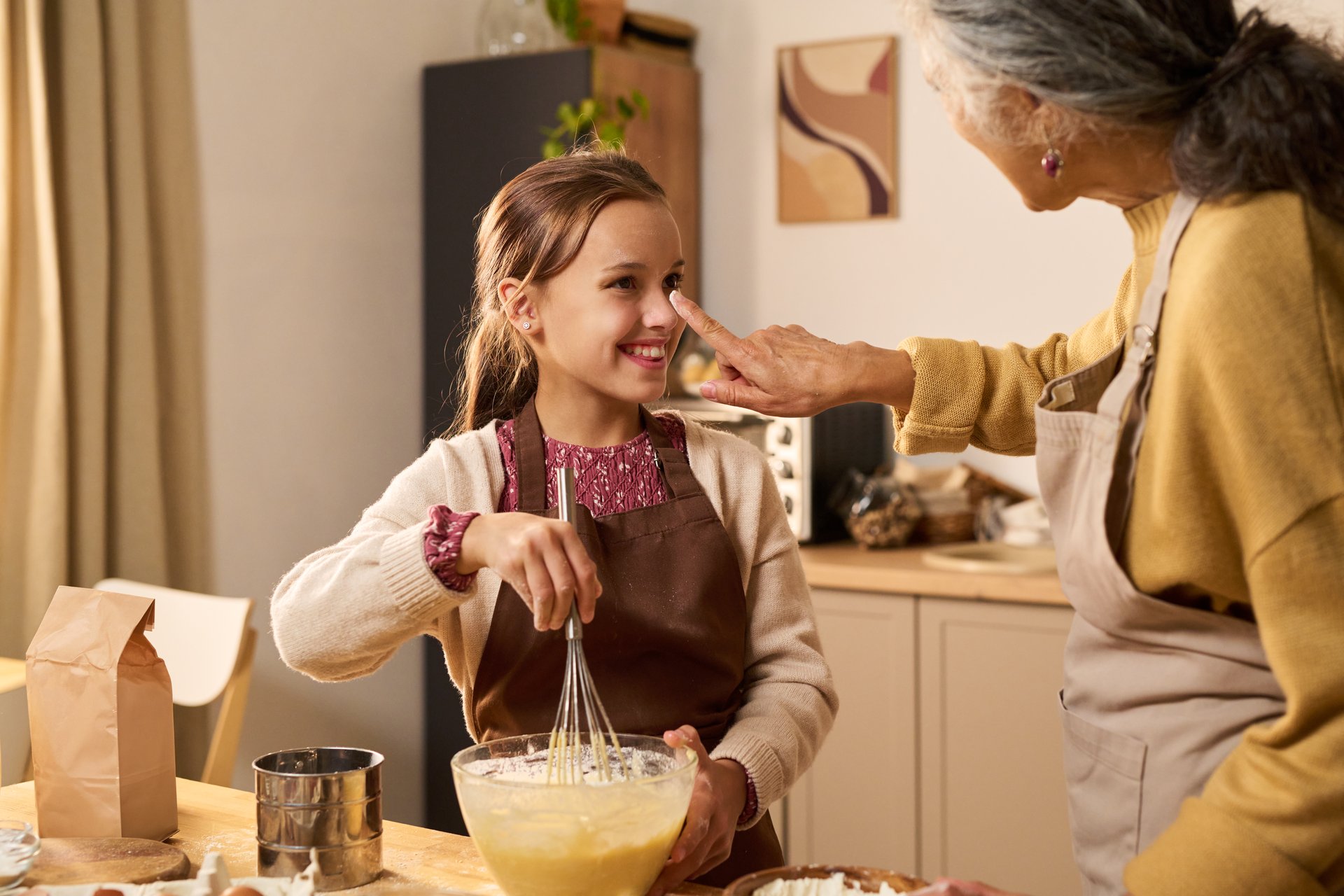 Caucasian girl smiling while whisking batter in kitchen, senior Caucasian woman touching her nose playfully, both wearing aprons and engaging in baking activity together