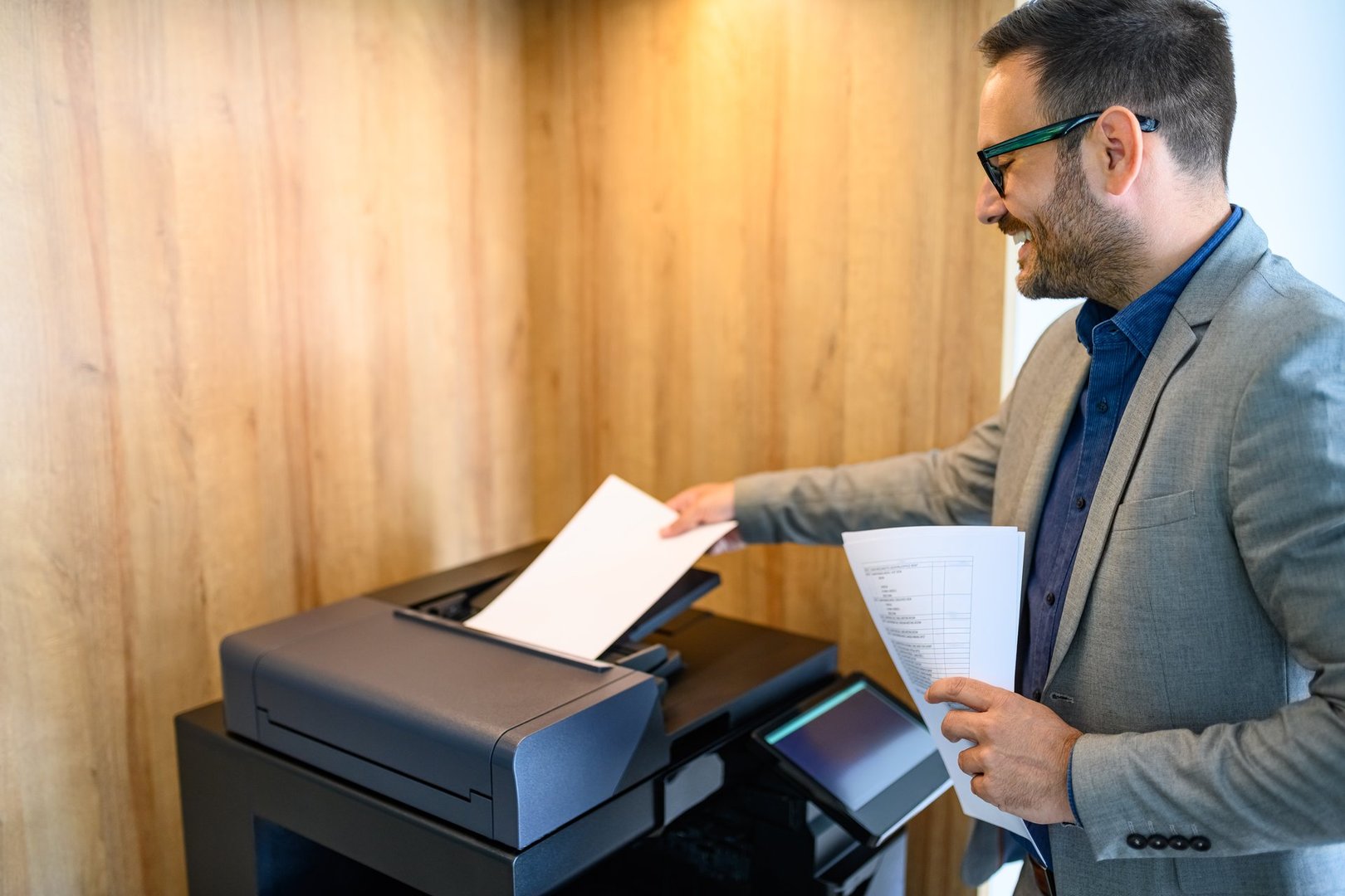 Side view of young smiling businessman putting papers into photocopier machine in modern office