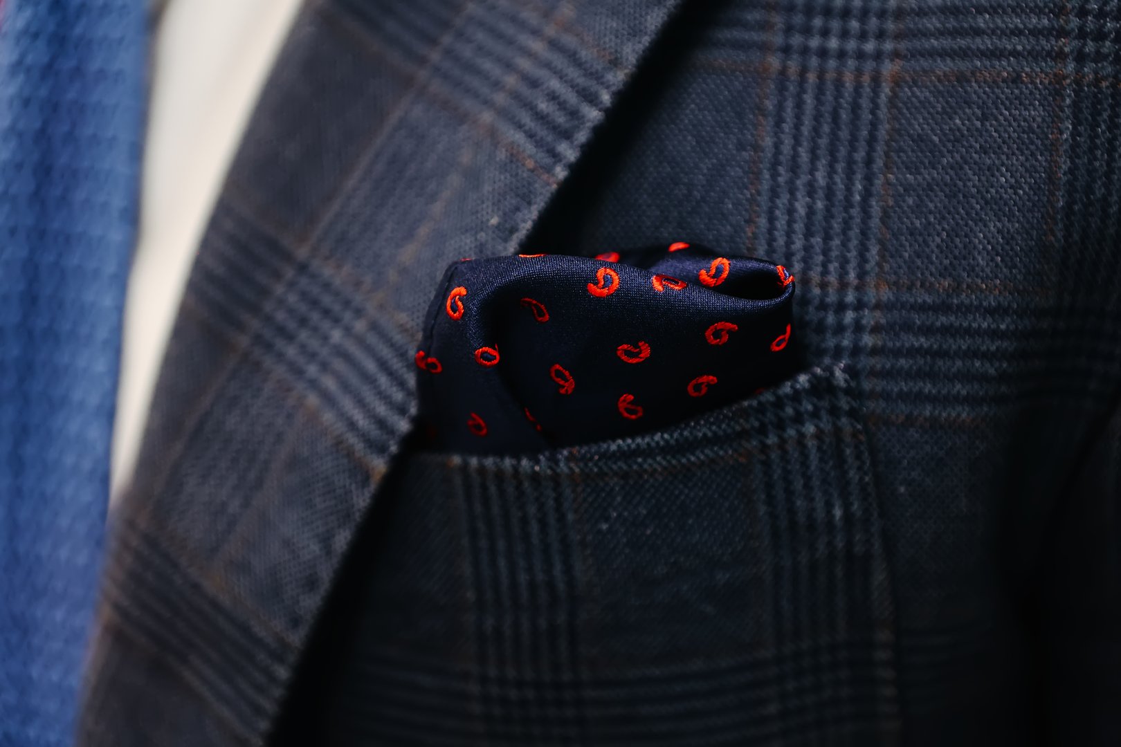 Close-up of a sophisticated navy blue pocket square with red paisley patterns elegantly tucked into the pocket of a plaid suit jacket, symbolizing refined style and sartorial elegance.