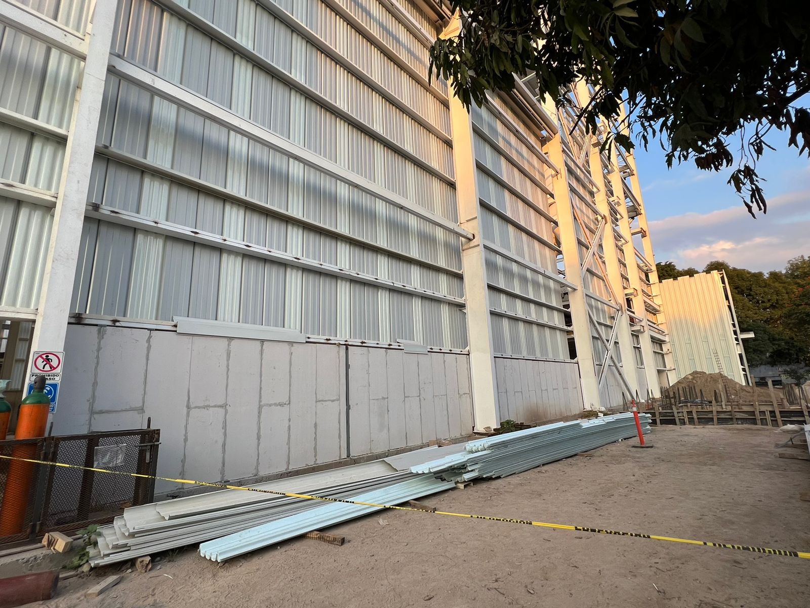 Construction site with a partially built metal structure, piled metal sheets, caution tape, and a no-smoking sign.