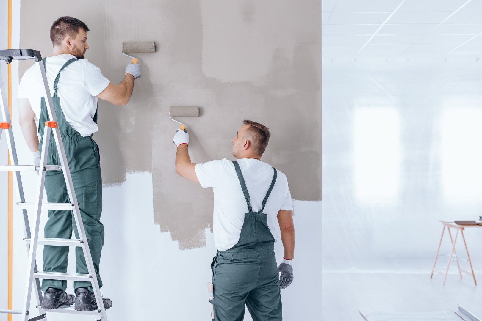 Man on ladder painting wall on beige color using roller next to painter in overalls