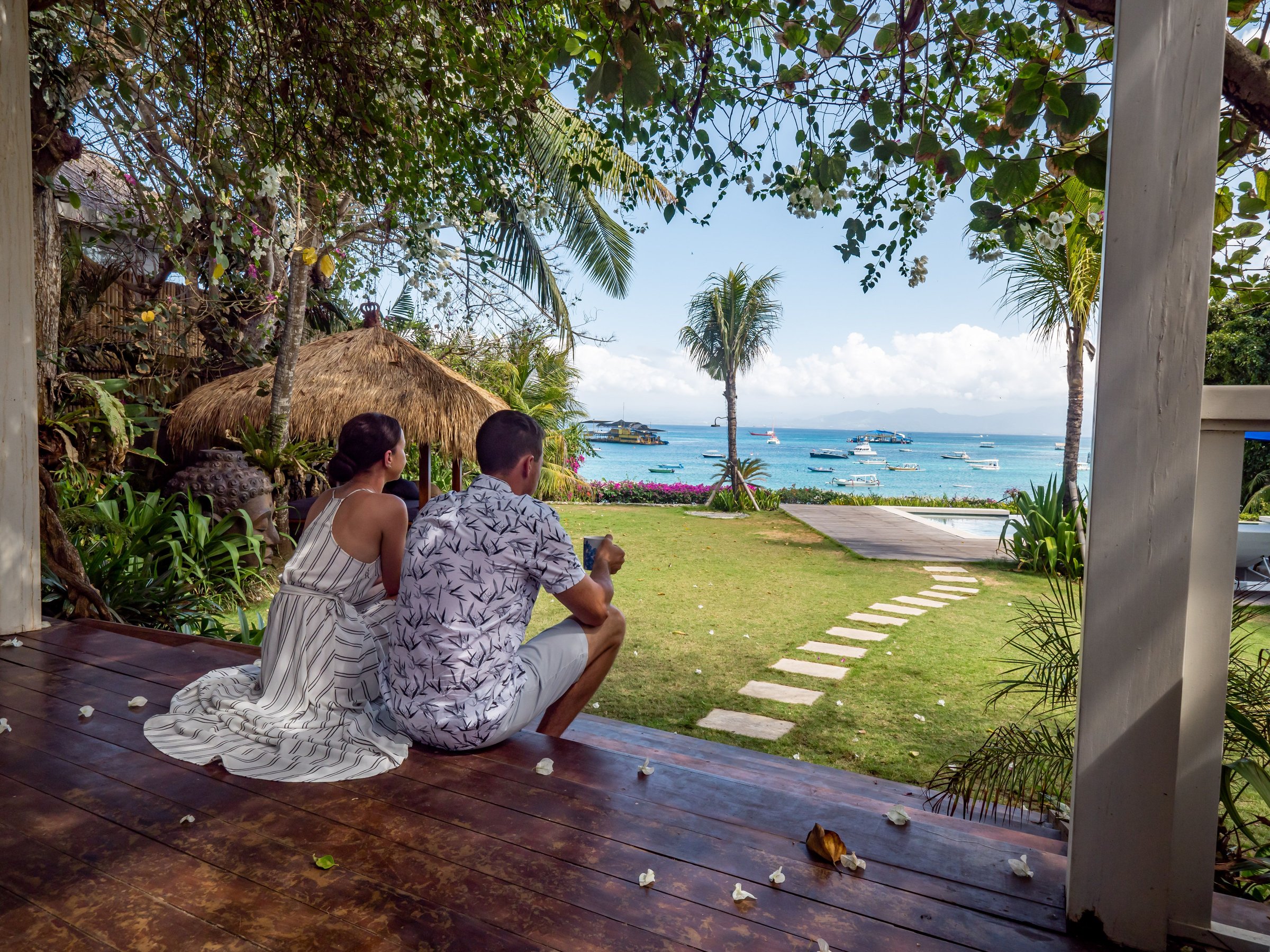 A couple enjoying a tropical beach view from a wooden deck, with lush greenery and boats in the distance.