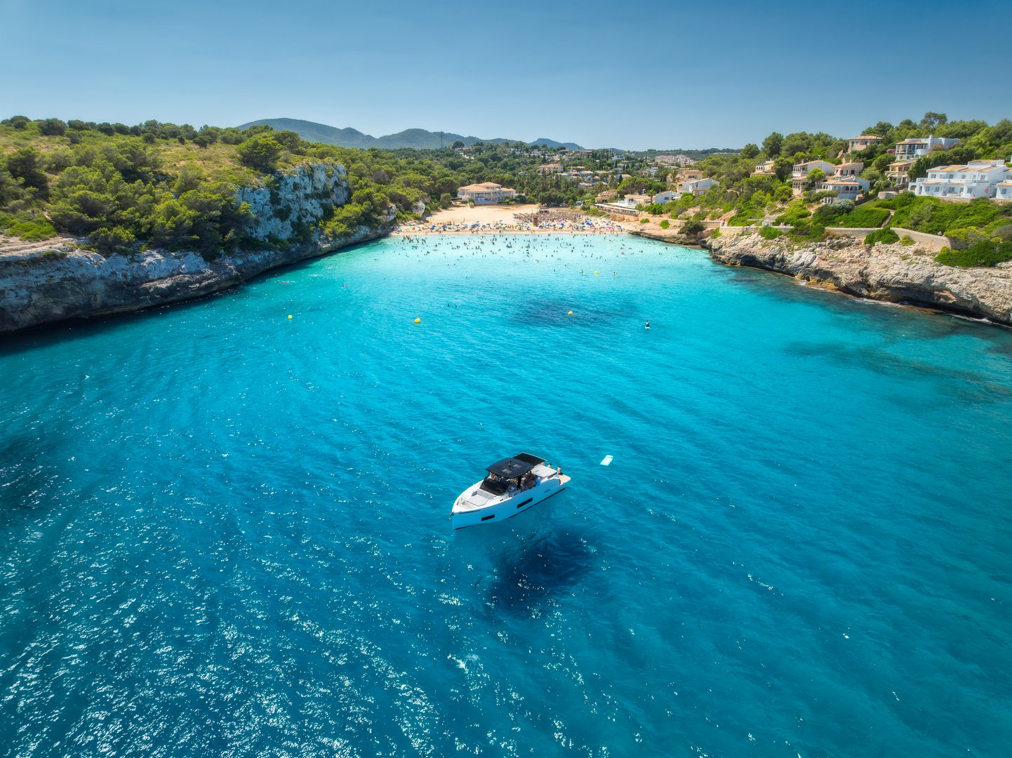 Aerial view of boat and serene sandy beach cove in Manacor, Balearic Islands. Mallorca, Spain. A solitary yacht floats on crystal-clear turquoise waters, surrounded by lush greenery and coastal villas