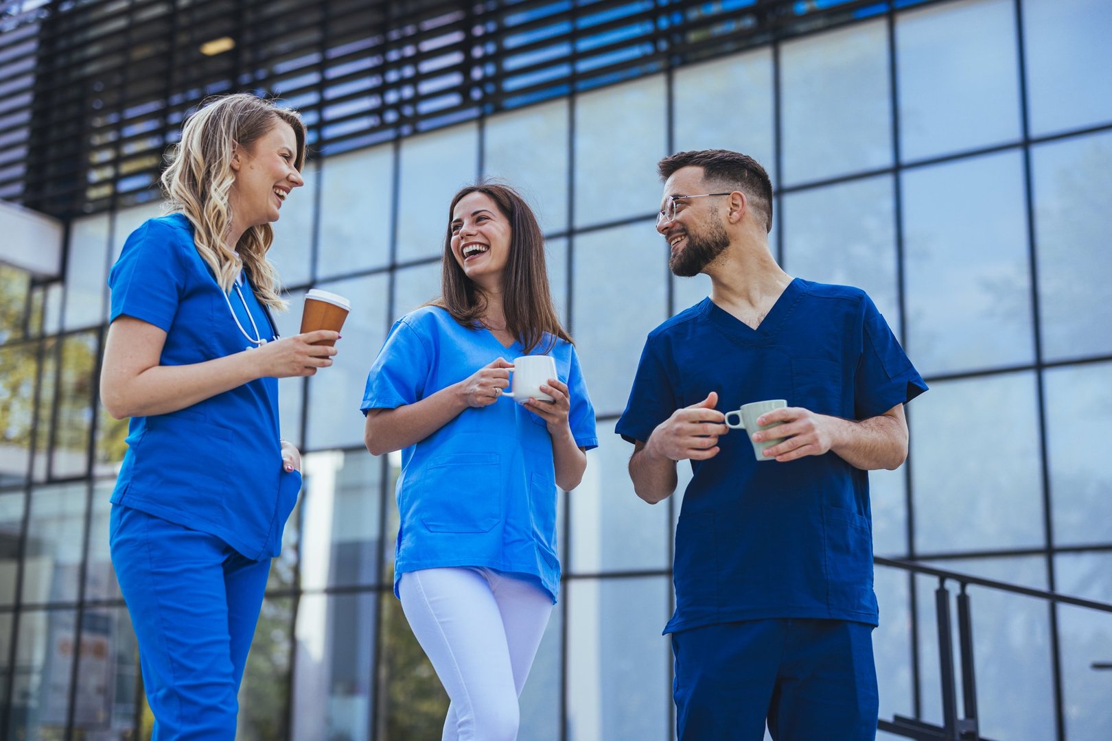 A group of healthcare professionals in uniforms smiling and talking while enjoying a coffee break outside a modern hospital building.