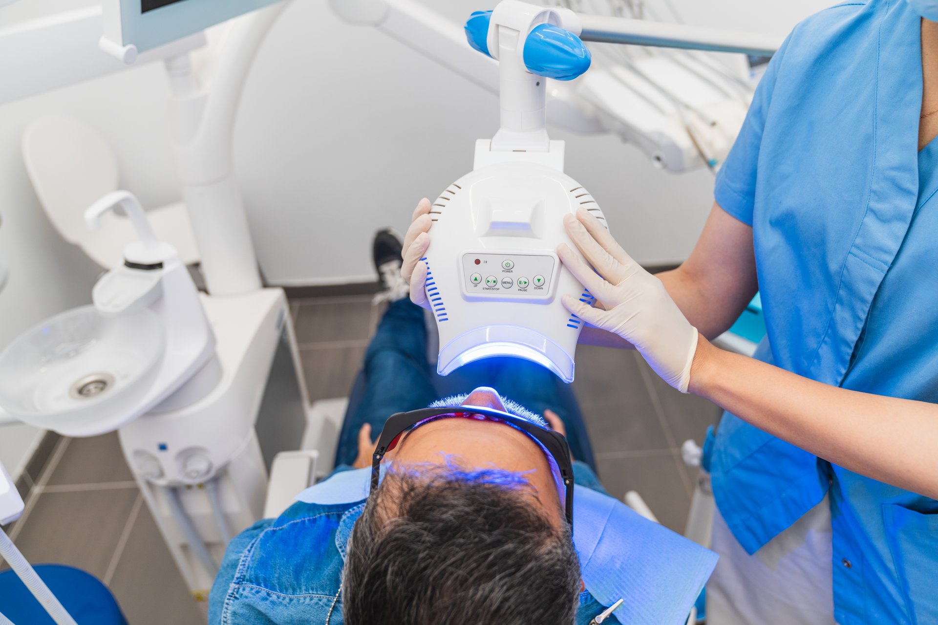 A man is getting his teeth whitened in a dentist's office. A woman is assisting him