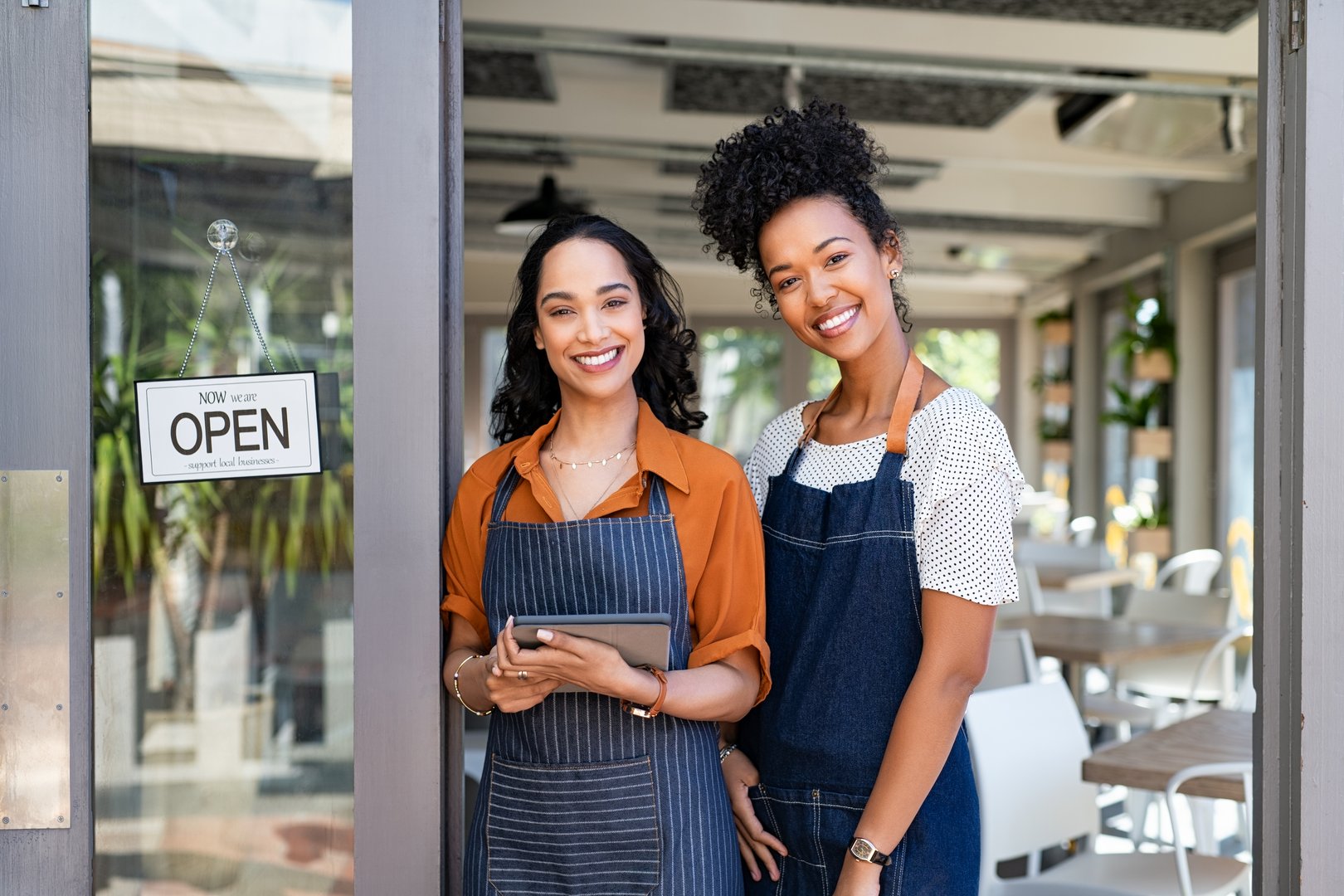 Portrait of smiling young latin woman holding digital tablet with black colleague at cafe entrance door. Two small business owners standing together at cafe entrance while smiling.