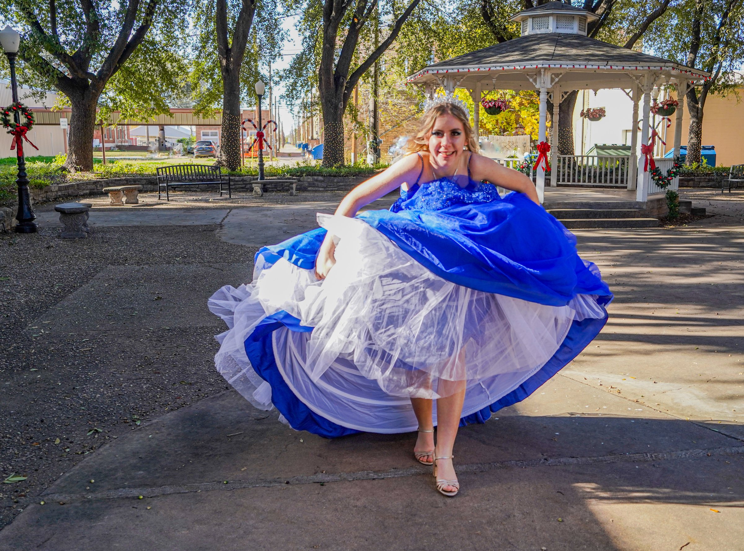 A playful photo of a teen girl, wearing a diamond tiara and diamond heart necklace, holding up her royal blue ball gown and crinoline as she runs in high heels. A gazebo decorated for Christmas is in the background.