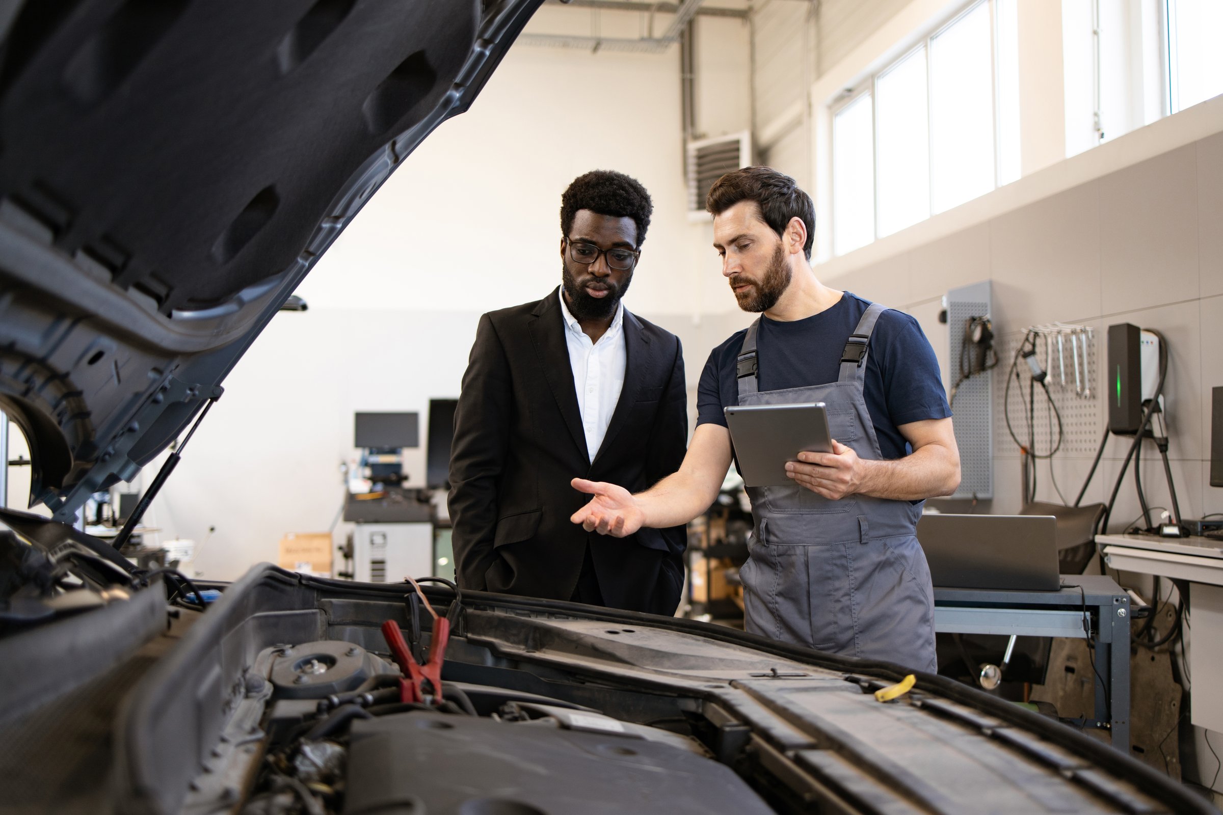 Mechanic in work overalls explaining car issues to customer in suit in modern auto repair shop. Professional consultation about vehicle maintenance and repair process.