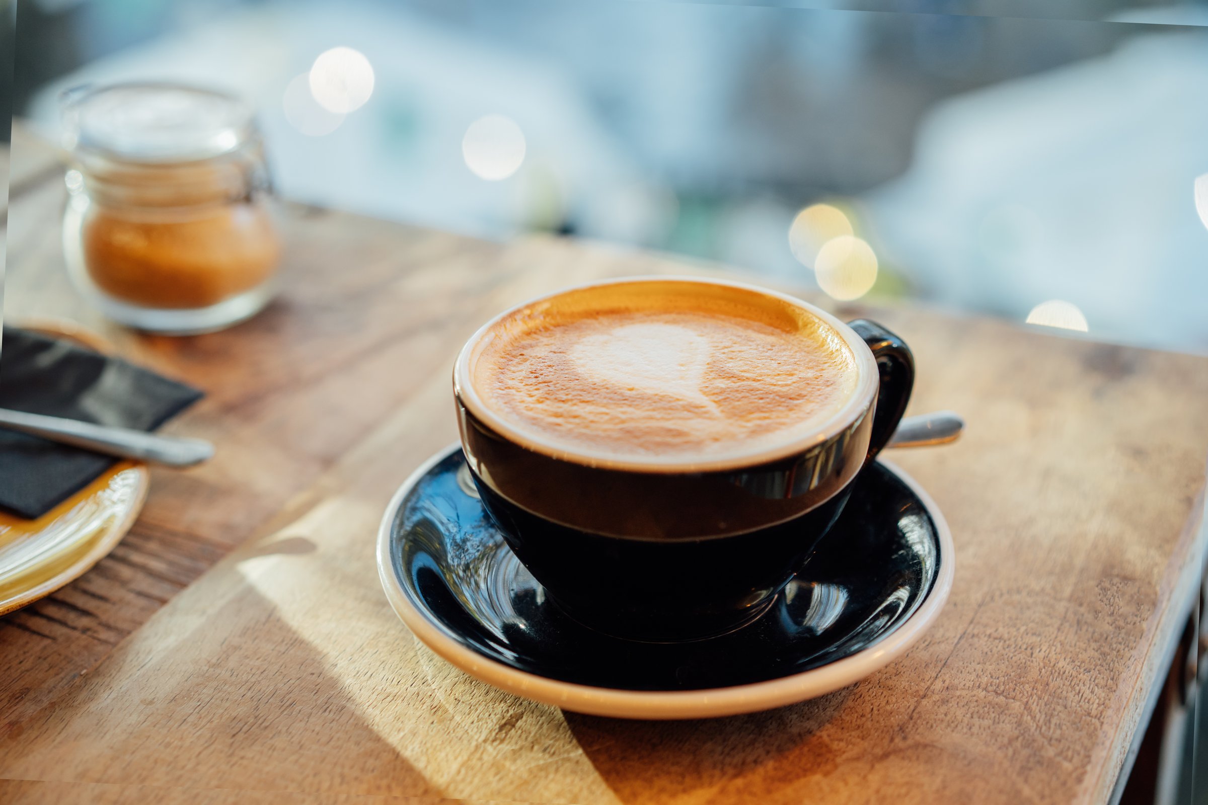 Close up hot cappuccino ceramic black coffee cup with heart shape latte art on the wooden craft table at cafe shop. Warm and cozy food and drink concept. Soft selective focus.