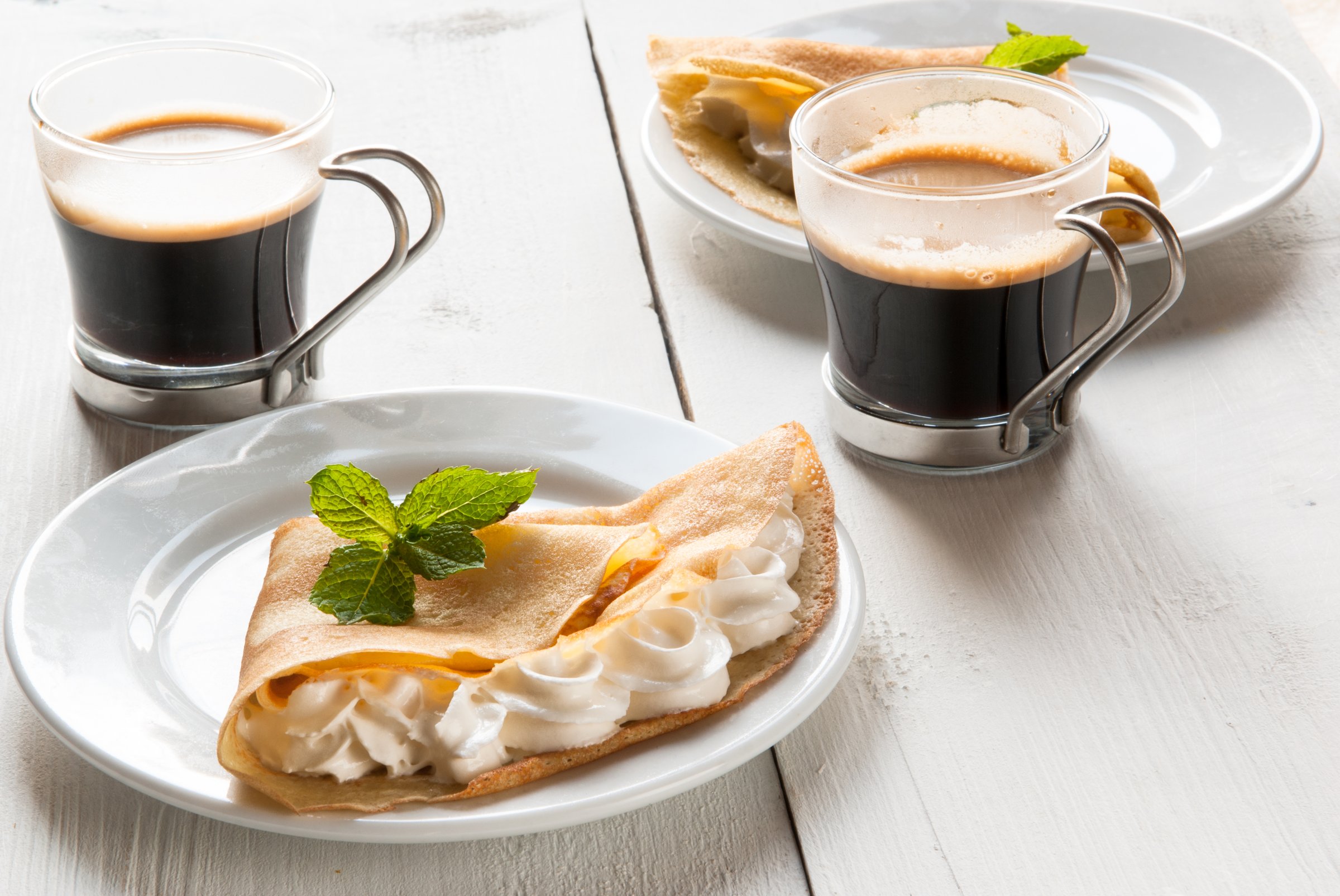 A light breakfast or lunch for two persons, with pancakes (crepes) and whipped cream. On the table is the coffee. Sunny morning, white wooden table