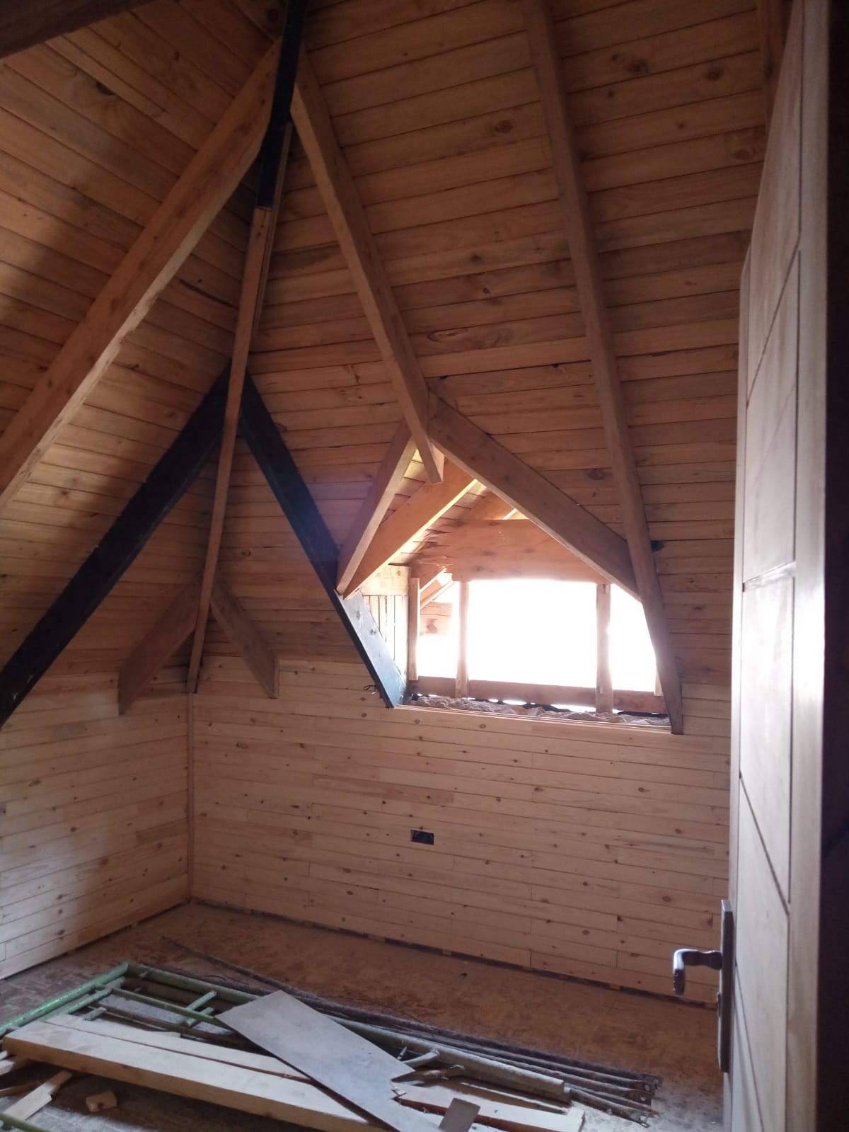 Unfinished attic with wooden paneling, exposed beams, a small window, and construction materials on the floor.