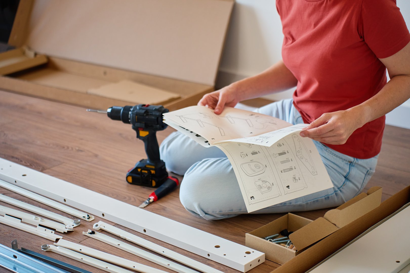 Woman sitting on floor reading instruction manual during furniture assembling. Person sitting on floor near tools and parts of new bed. Concept of DIY home improvement and furniture installation