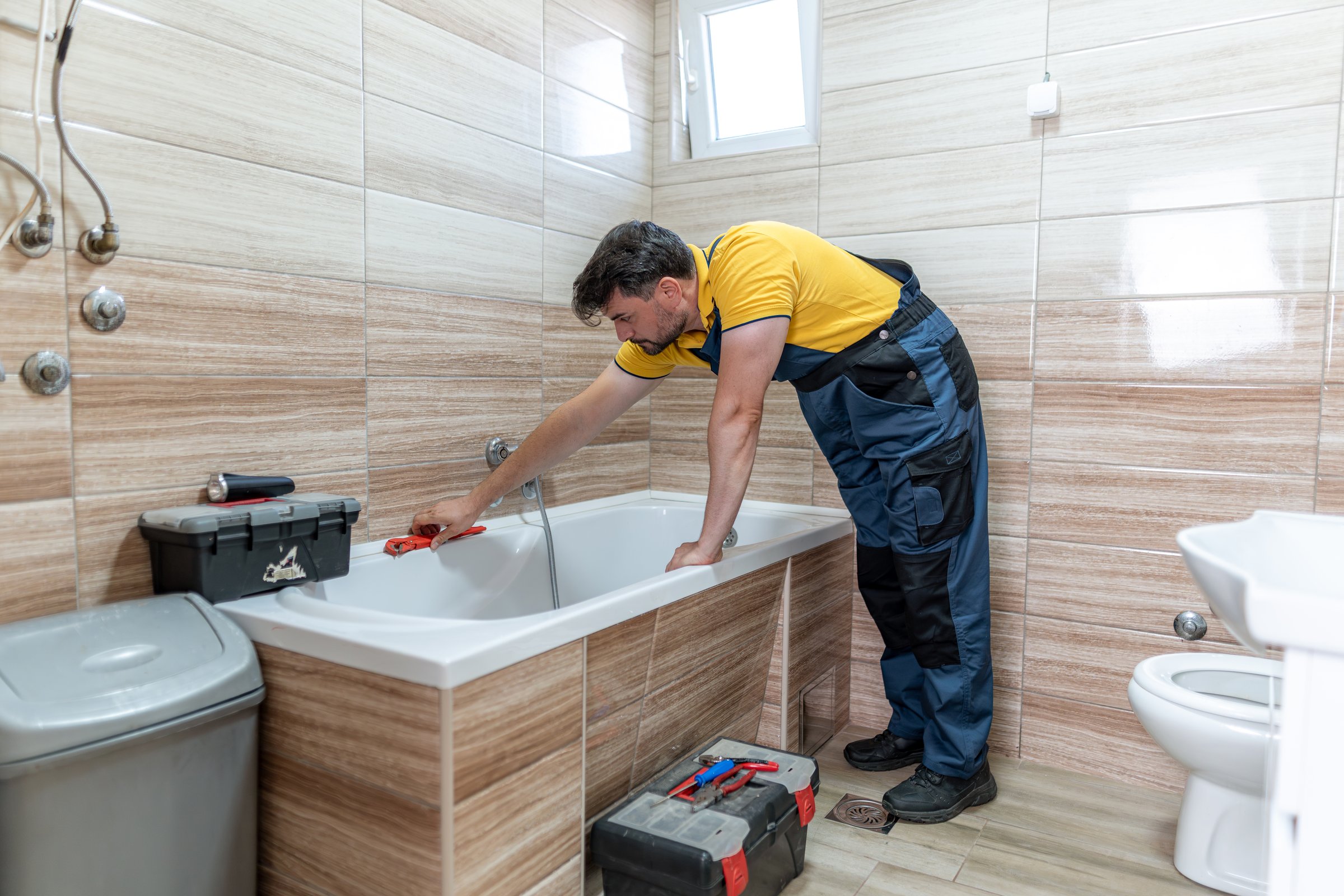 A skilled plumber is working on a bathtub in a modern bathroom. The space features light-colored tiles and essential plumbing tools are visible nearby. The professional focuses on the plumbing issue.