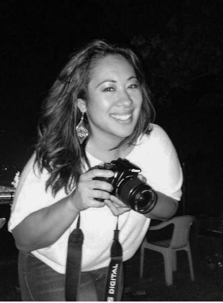 Black and white image of a woman smiling, holding a camera, with long hair and wearing a white top.