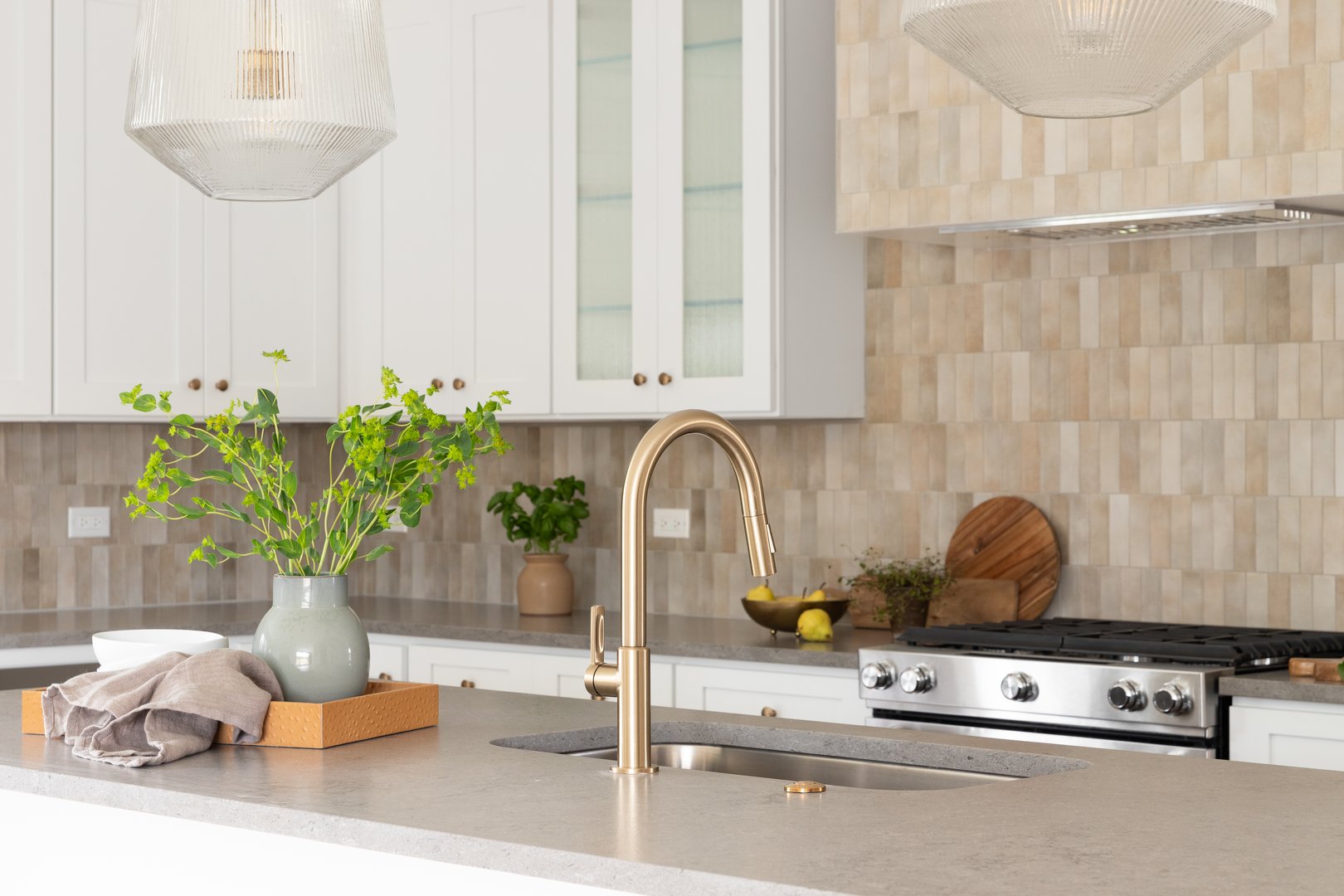 A kitchen faucet detail with a grey stone countertop island, gold faucet, stainless steel appliances, brown tile backsplash, and white cabinets.