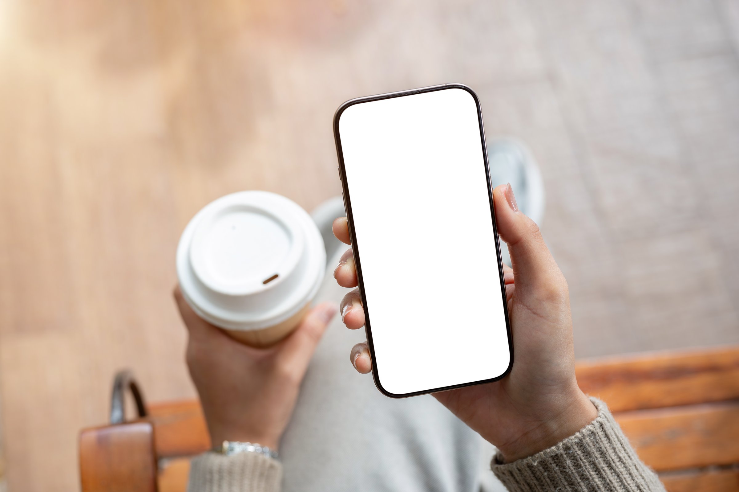 A top view of a smartphone with a white screen mockup in a woman's hand. A woman holds a coffee cup and a smartphone. people, lifestyle, wireless technology