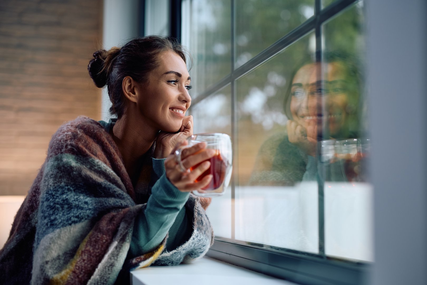 Young smiling woman drinking tea and looking through the window at home. Copy space.