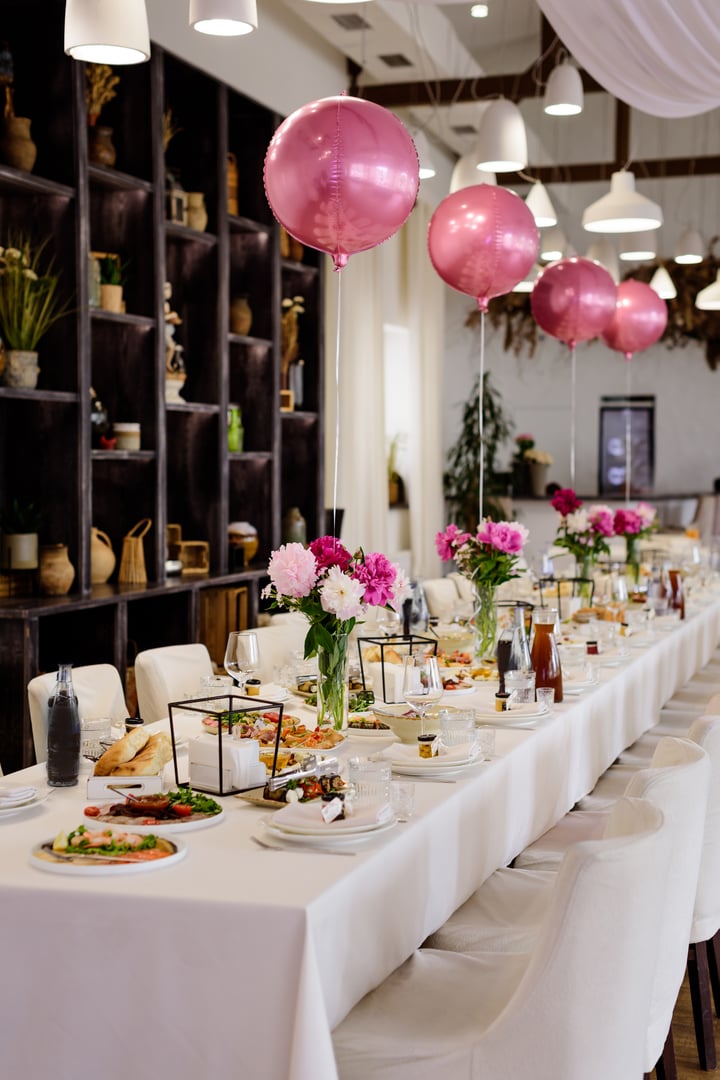 Elegant long banquet table set for a celebration, adorned with vibrant pink and white peony centerpieces, ambient candlelight, and festive pink balloons, creating a luxurious and inviting atmosphere