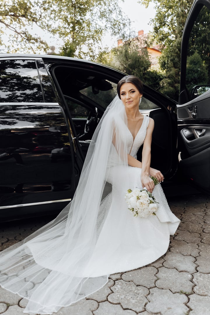 A bride is sitting in the back of a black car with a white veil. She is holding a bouquet of flowers