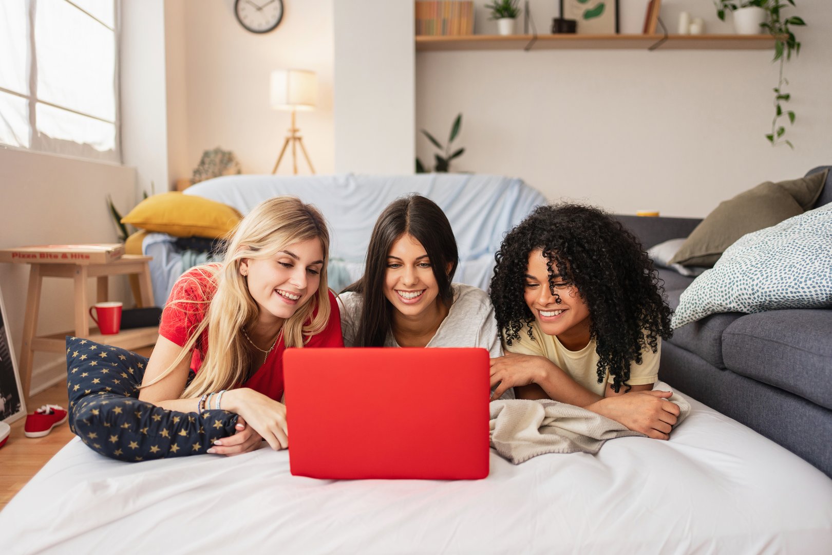 Three female flatmate friends watching films on the computer, laughing and relaxing in bed
