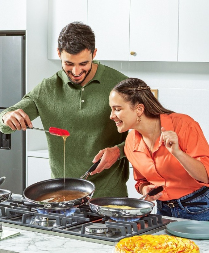 Two people cooking together, smiling, as one pours sauce from a spoon into a pan on a kitchen stovetop.