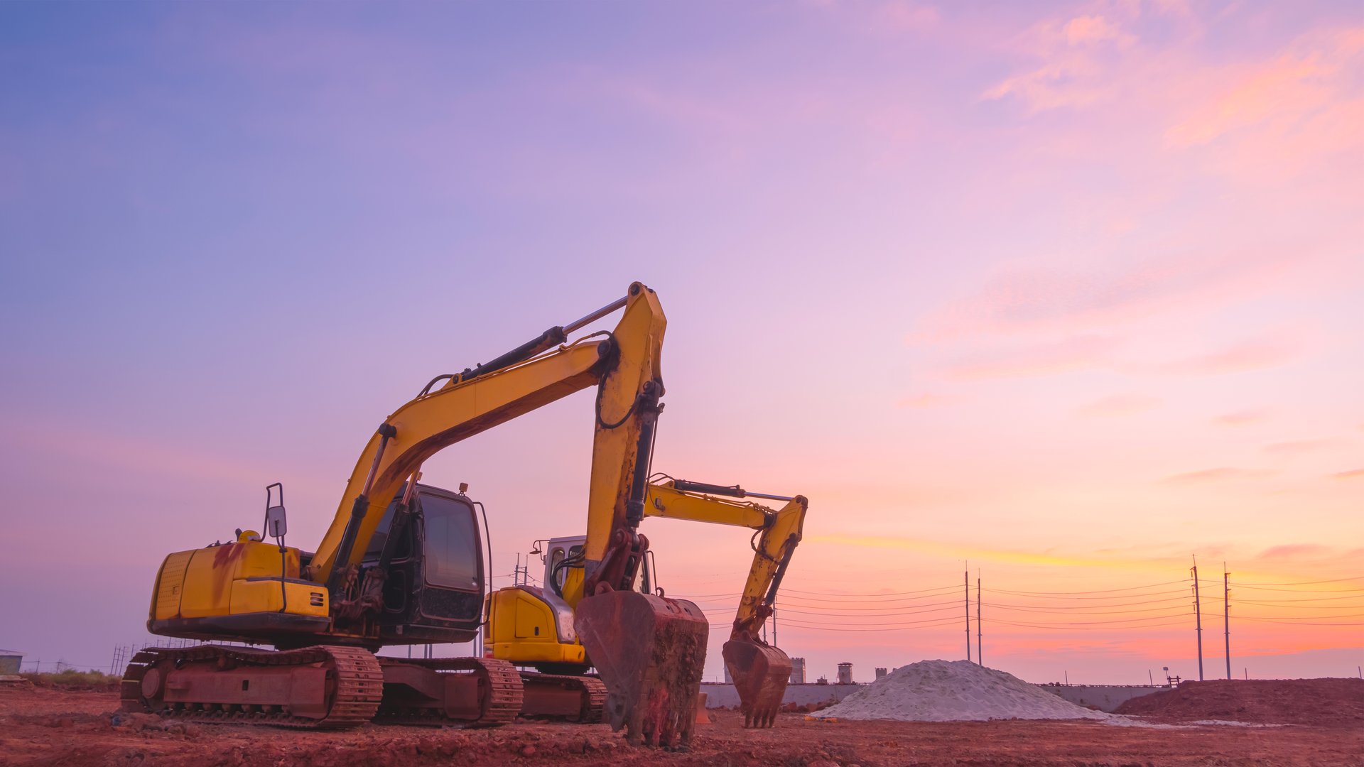 Silhouette of 2 excavators parked after work in land filling area of new industrial factory construction site against sunset sky background