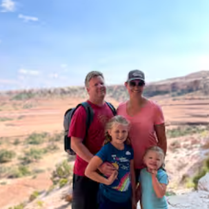 Family of four posing in a desert landscape, with rocky terrain and clear blue sky in the background.